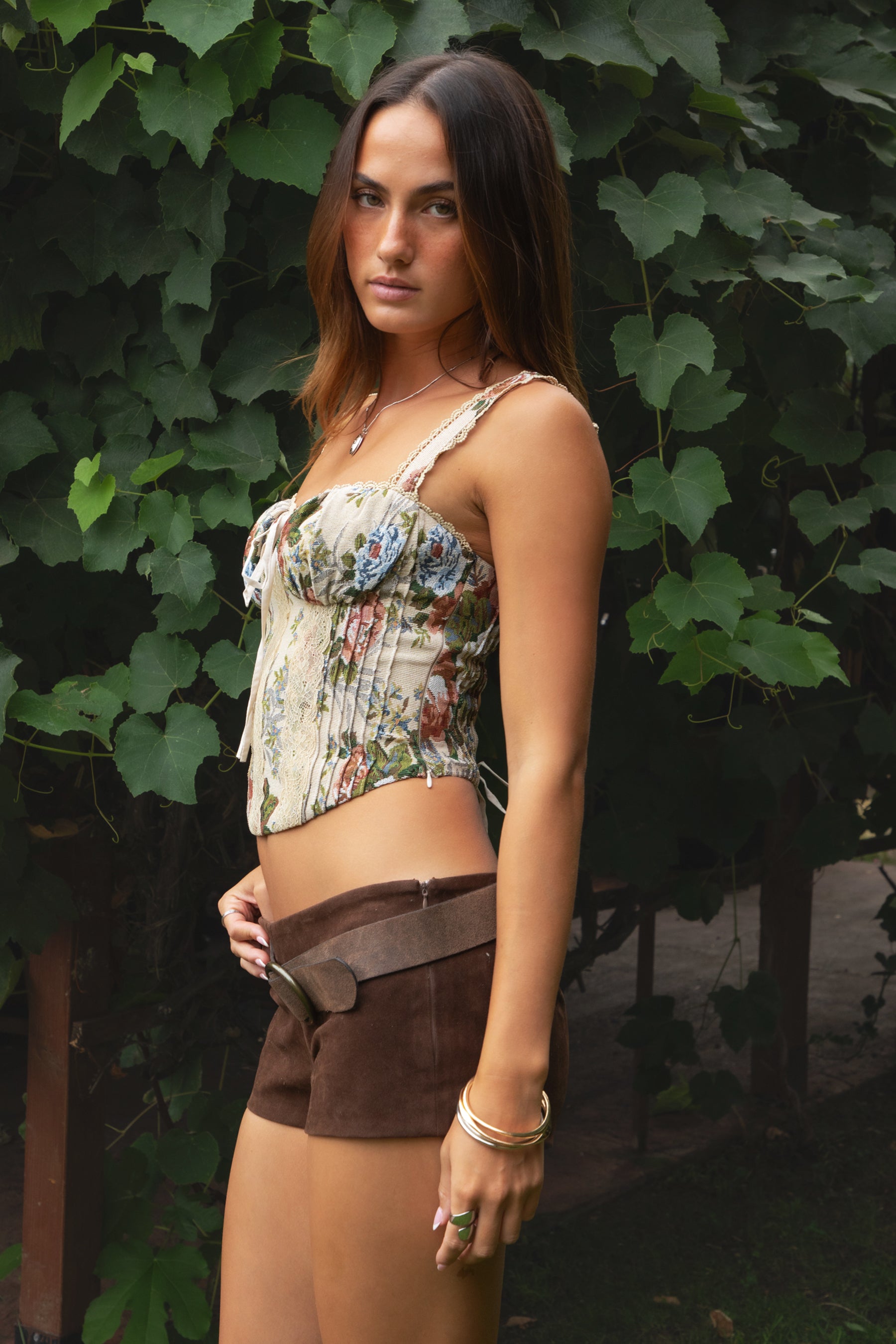 Woman wearing the TIC TOC TA31127 BEYLA floral tapestry lace bustier corset top with lace straps, bow detail, and lace-up back paired with brown suede shorts in front of greenery. 