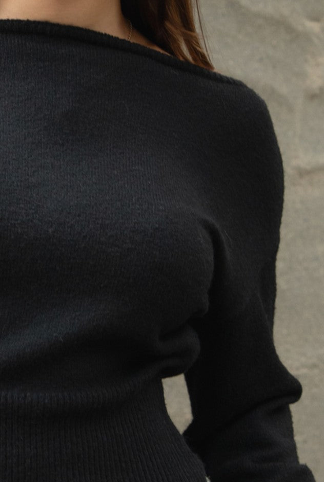 Woman wearing TIC TOC TA31843 AURELLE black off the shoulder knit sweater top with dropped shoulders and relaxed long sleeves paired with a pleated brown mini skirt in an indoor setting.