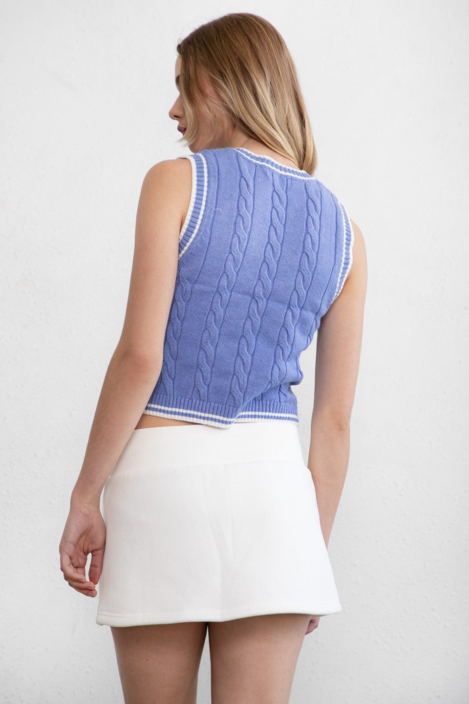 Woman wearing TIC TOC TC29858 WESTBRIDGE cable knit chunky vest with v neck and semi cropped length in classic ribbed hem design in color blue paired witha white mini skirt. 