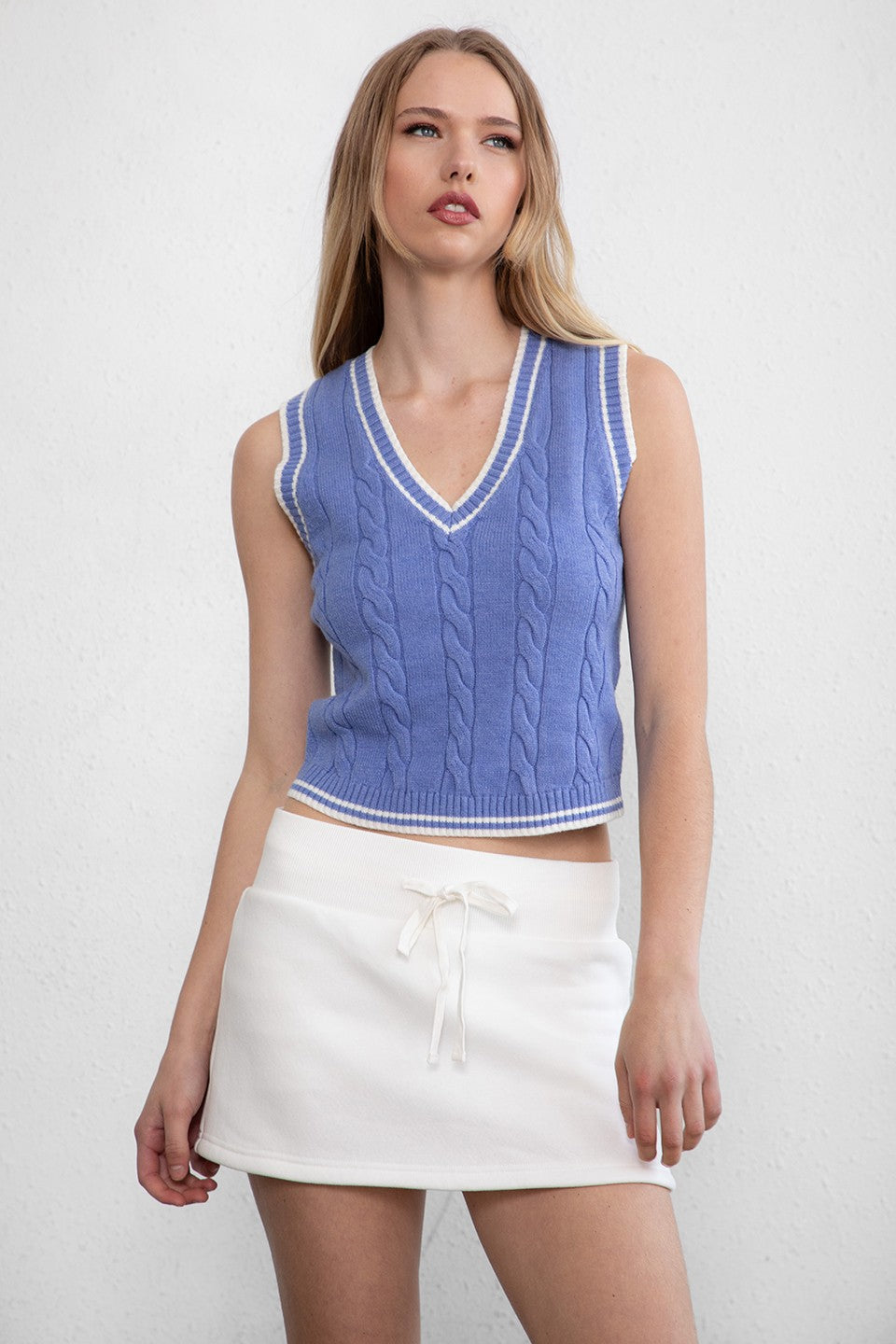 Woman wearing TIC TOC TC29858 WESTBRIDGE cable knit chunky vest with v neck and semi cropped length in classic ribbed hem design in color blue paired witha white mini skirt. 
