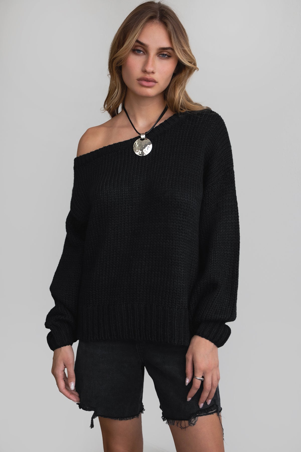 Woman wearing TIC TOC TC30554 BRANDY ribbed knit oversized boat neck off the shoulder sweater in black with denim shorts. 