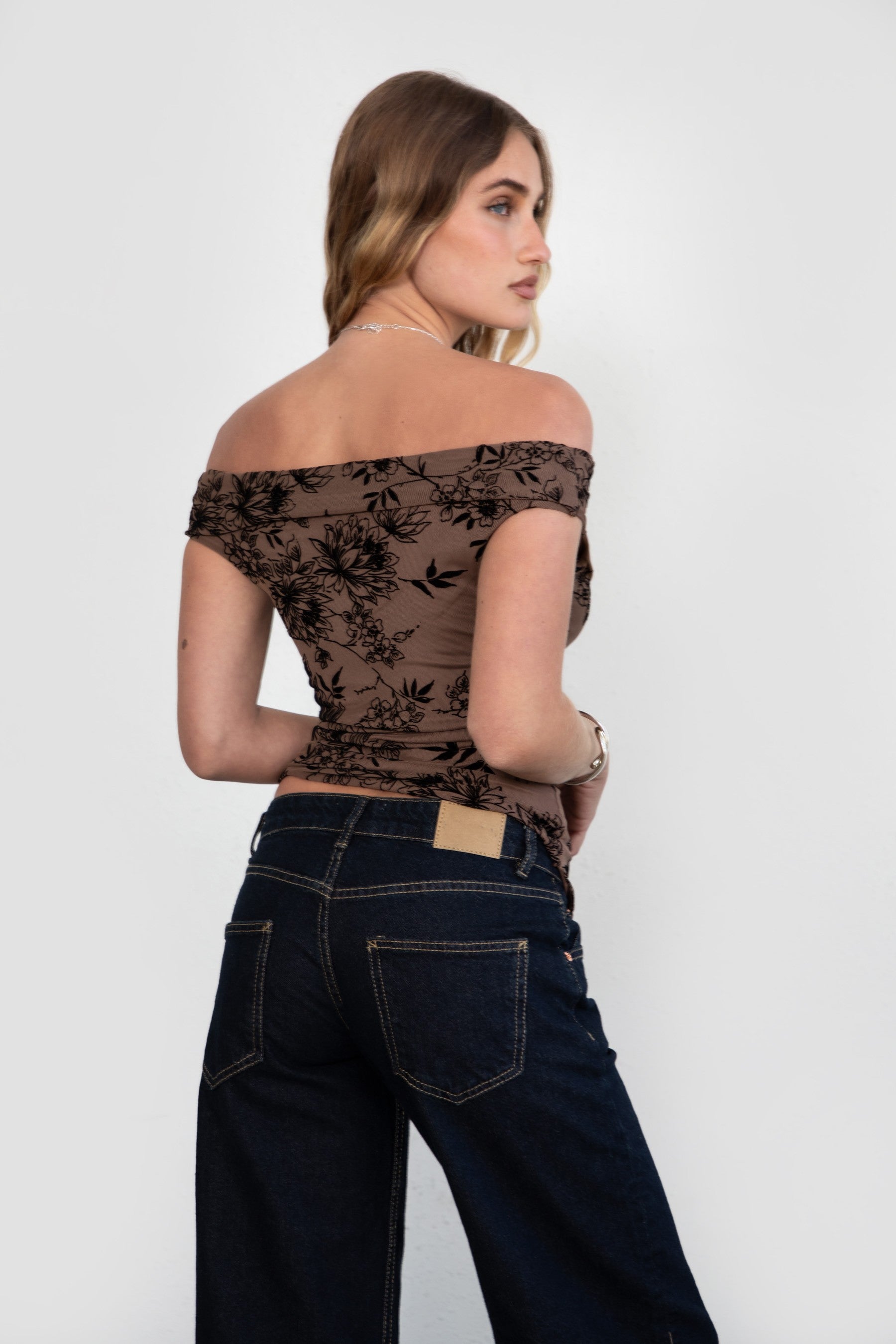 Woman wearing a black floral off-shoulder top and dark jeans on a white background
