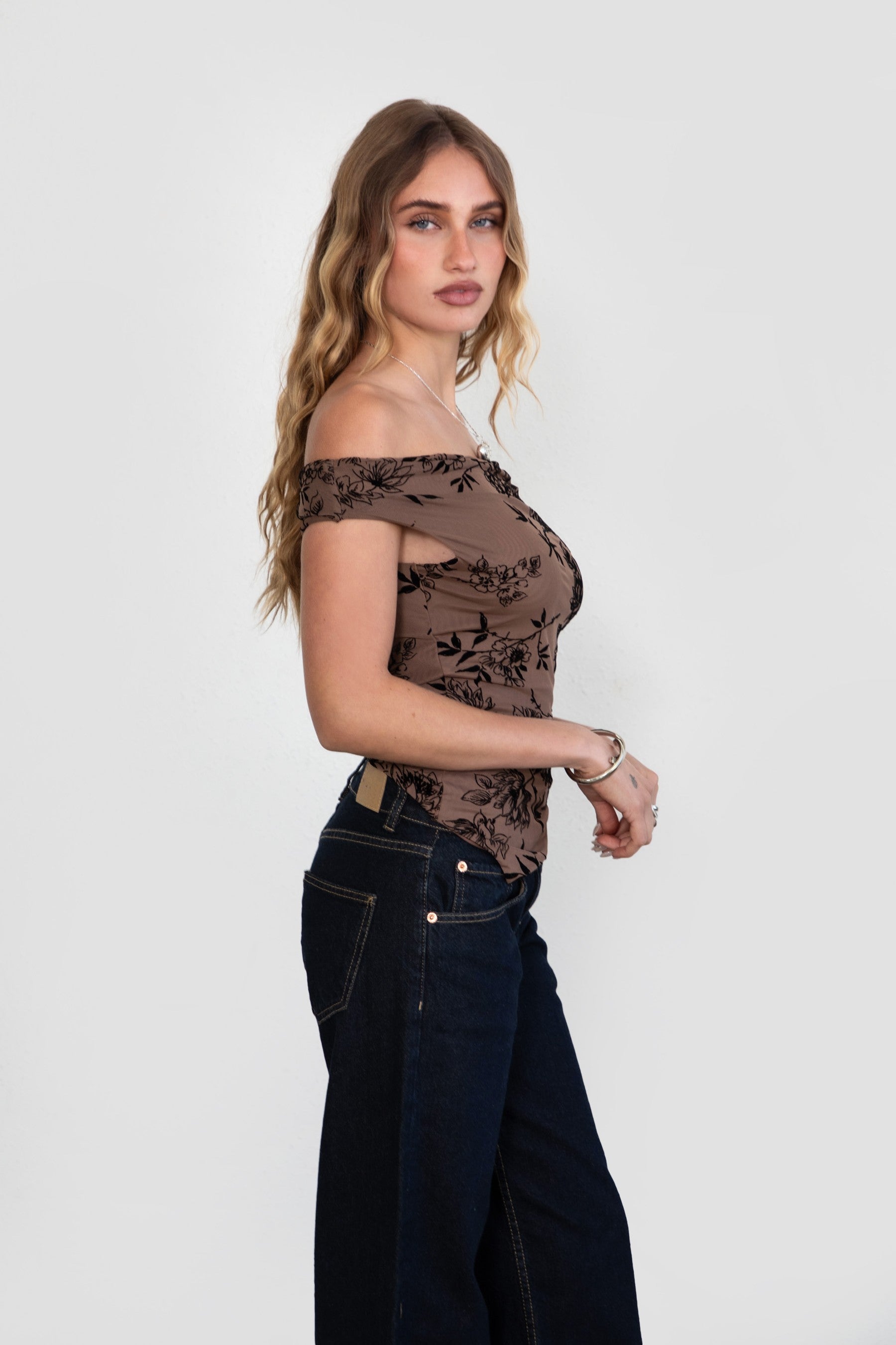 Woman wearing a brown floral off-shoulder top and dark jeans on a white background