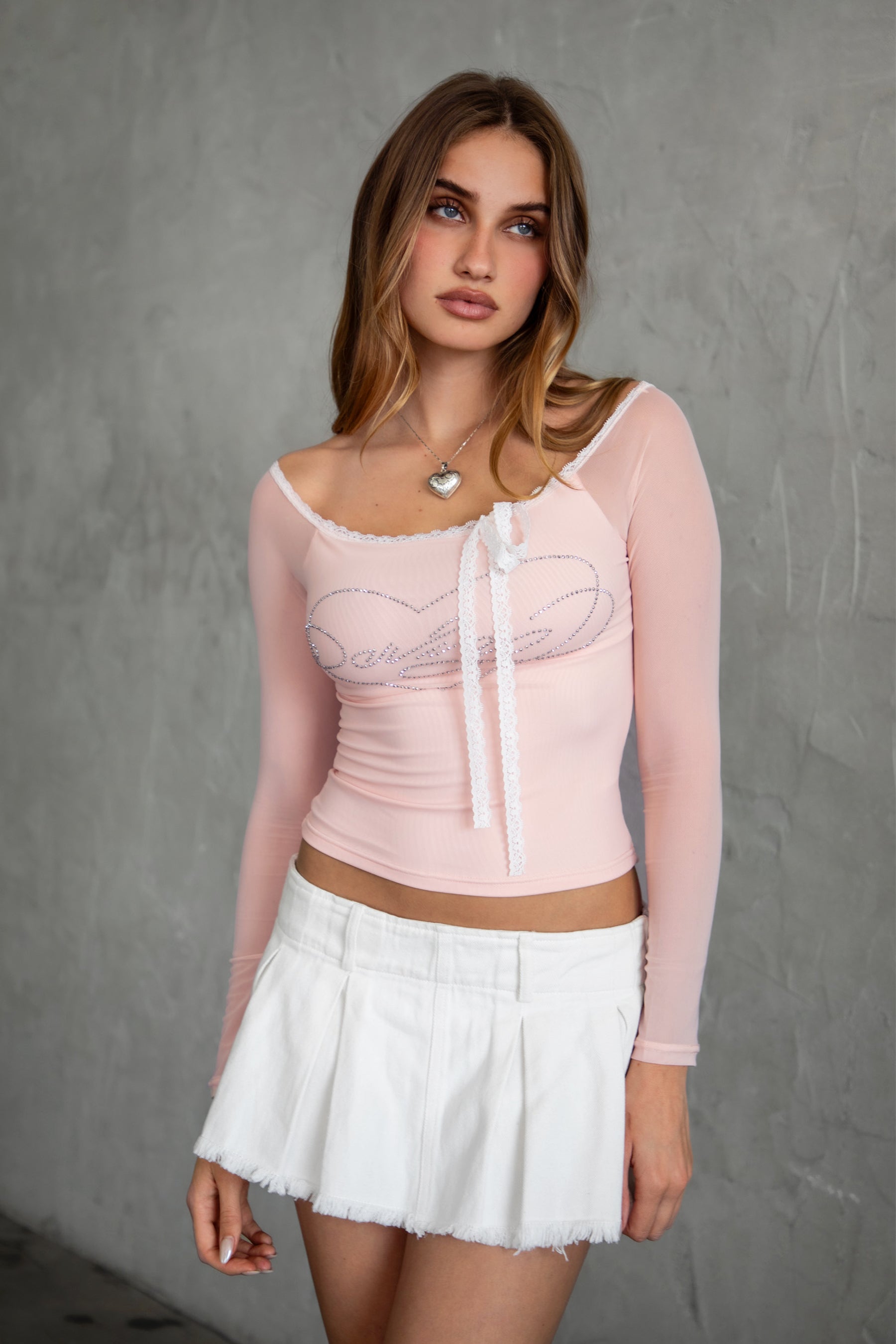 Woman wearing TIC TOC TC31111 ADORA mesh rhinestone long sleeve top with off the shoulder neckline and lace trim tie detail paired with a white denim mini skirt. 