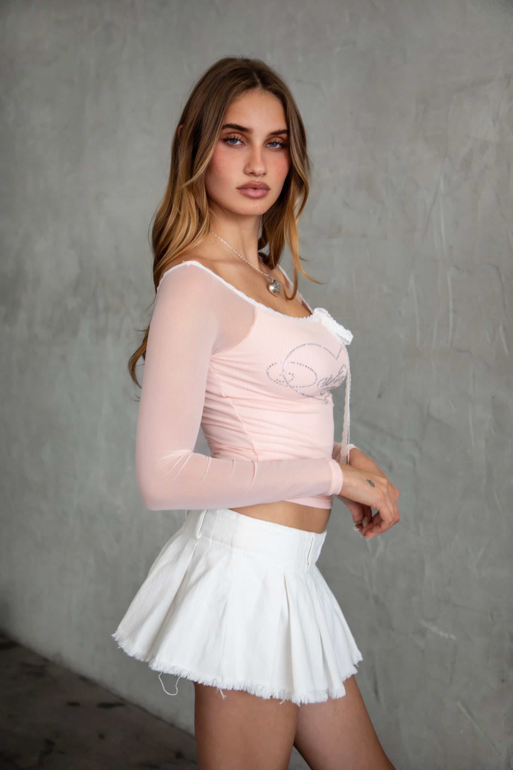 Woman wearing TIC TOC TC31111 ADORA mesh rhinestone long sleeve top with off the shoulder neckline and lace trim tie detail paired with a white denim mini skirt. 
