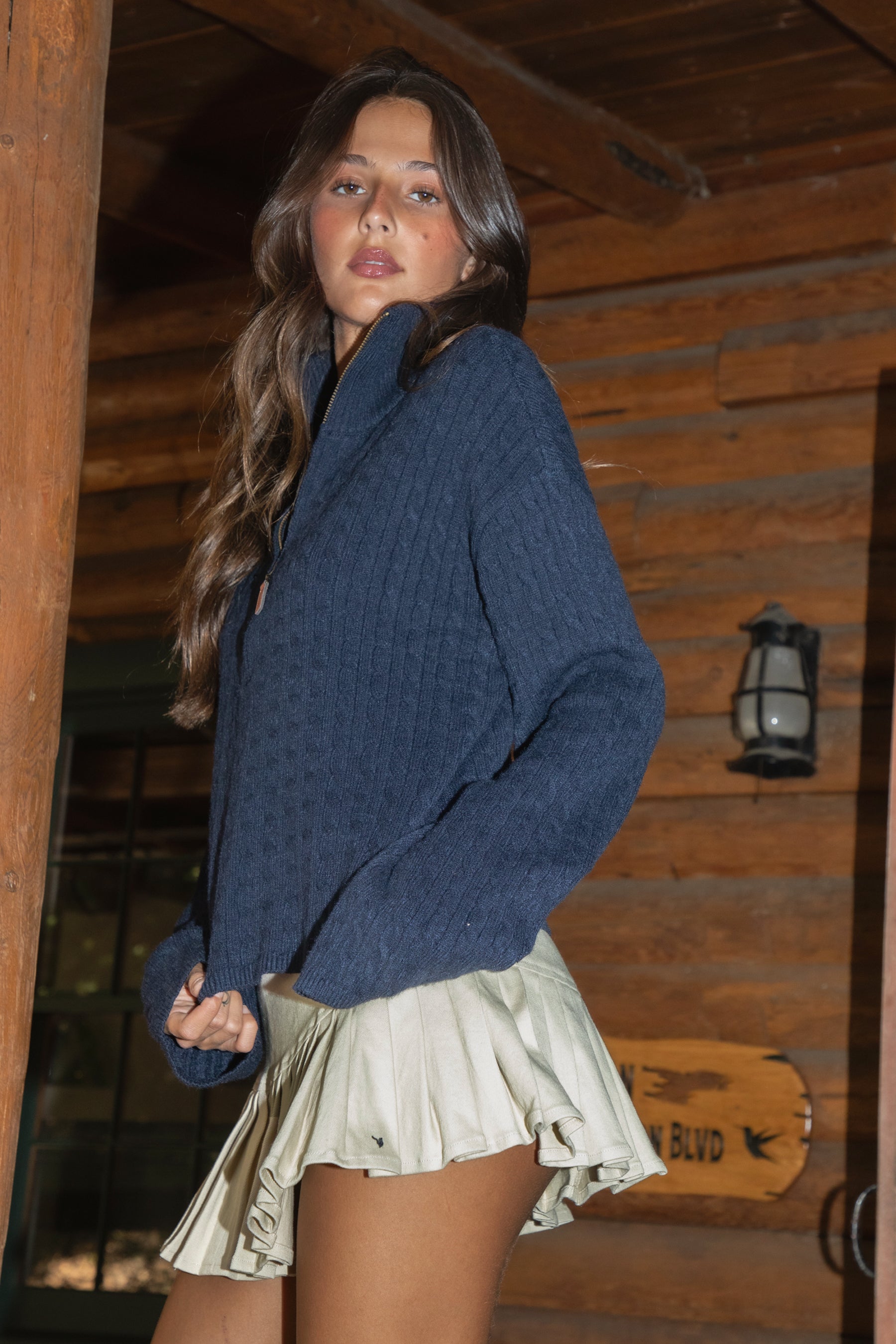 A woman wearing the TIC TOC TC31470 FRANKIE cable knit sweater, featuring a half-zip front, mock neckline, and relaxed drop-shoulder fit, stands against a wooden wall with a beige skirt.