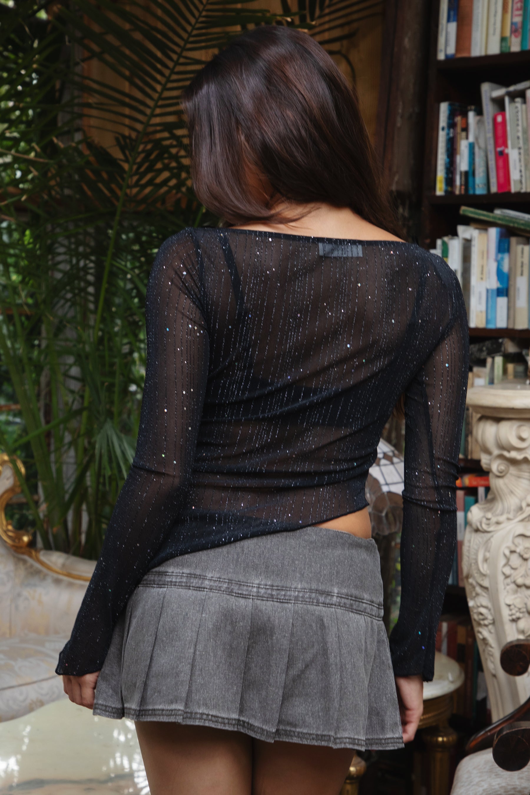 Woman wearing TIC TOC TC31640 MILLENIA lurex jersey long sleeve top with wide boat neckline and asymmetrical hem in sheer stretch fabric in color black, paired with a pleated denim mini skirt indoors. 