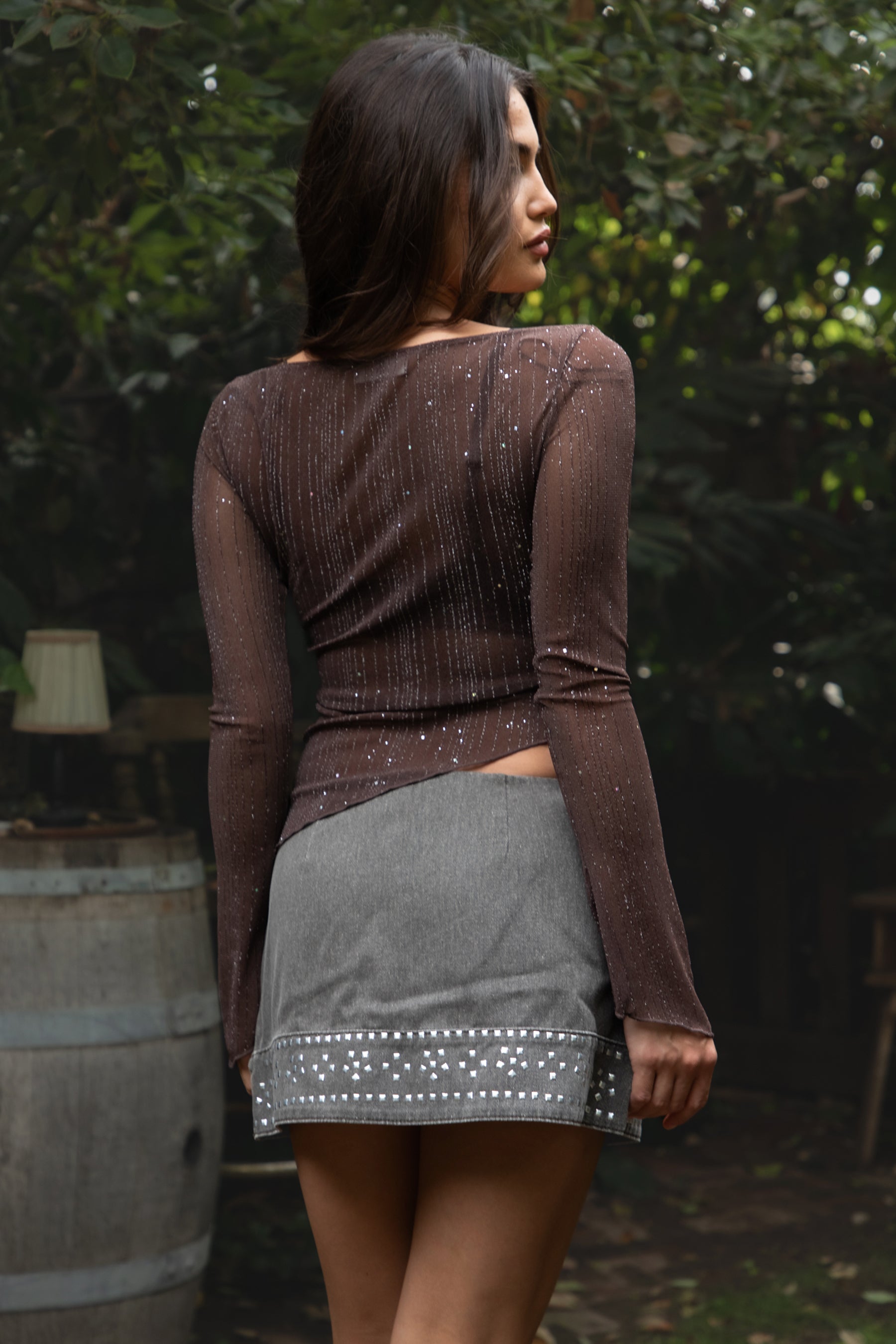 Woman wearing TIC TOC TC31640 MILLENIA lurex jersey long sleeve top with wide boat neckline and asymmetrical hem in sheer stretch fabric in color brown, paired with a denim mini skirt outdoors. 
