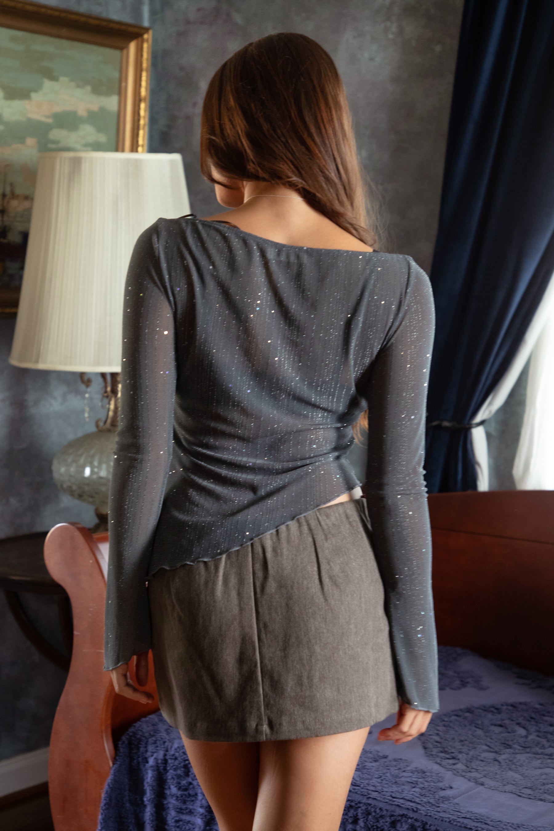Woman wearing TIC TOC TC31640 MILLENIA lurex jersey long sleeve top with wide boat neckline and asymmetrical hem in sheer stretch fabric in color grey, paired with a denim mini skirt indoors. 