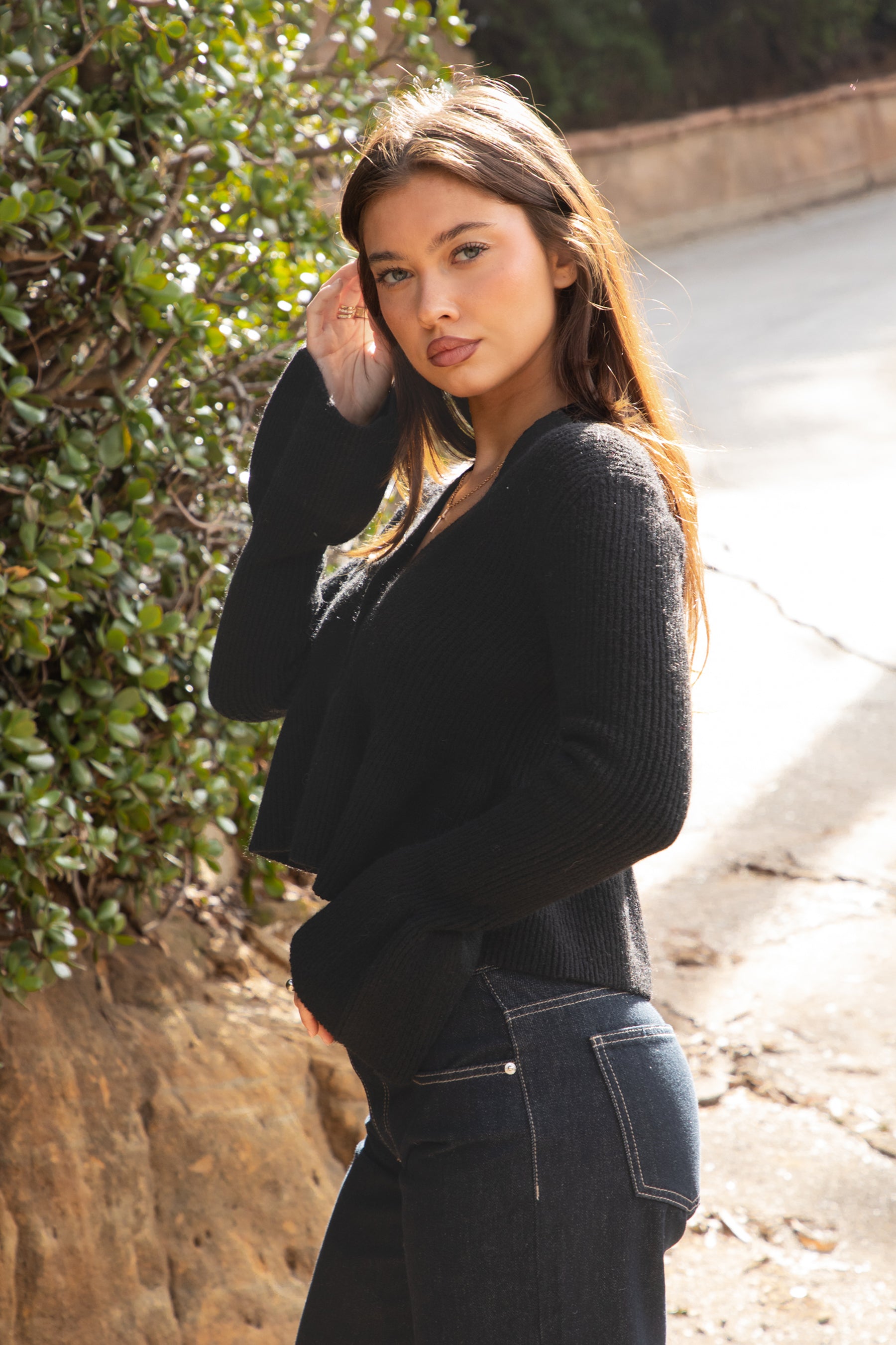 Woman wearing TIC TOC TC31678 LETTIE black sweater knit V-neck cardigan with flared sleeves, cropped peplum hem, and ribbed stretch fabric with dark denim jeans in an outdoor setting. 