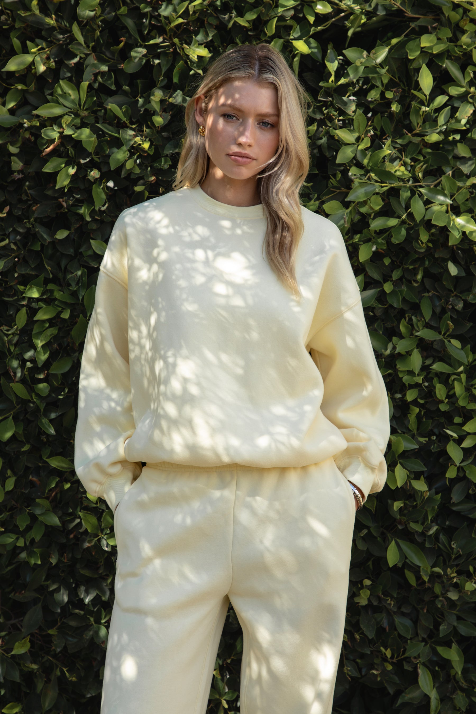 Woman wearing the TIC TOC TH28243 VARSITY pale yellow fleece crew neck pullover sweatshirt featuring dropped shoulders, ribbed trim, and plush fleece fabric with the matching bottoms standing against a green hedge.