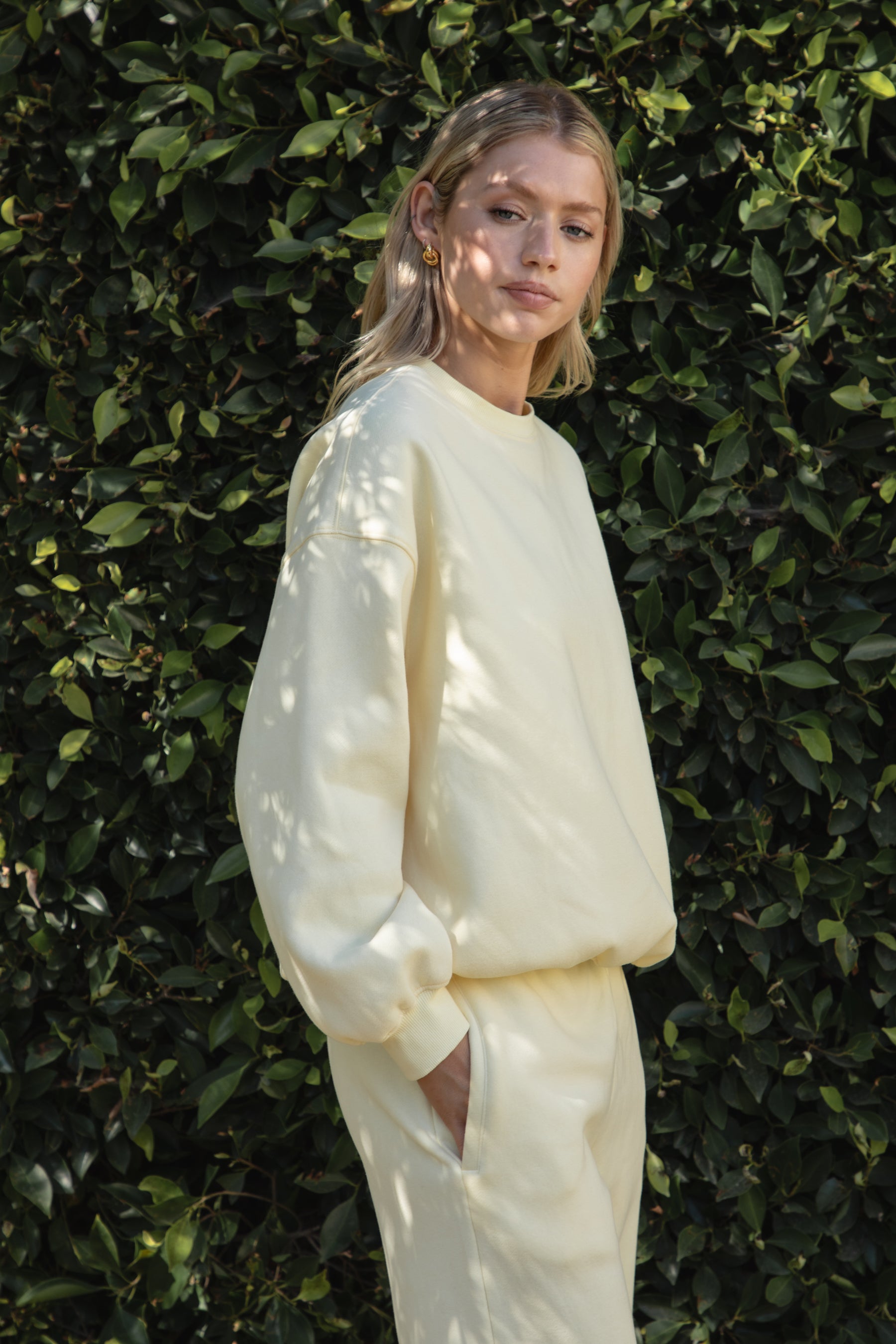Woman wearing the TIC TOC TH28243 VARSITY pale yellow fleece crew neck pullover sweatshirt featuring dropped shoulders, ribbed trim, and plush fleece fabric with the matching bottoms standing against a green hedge.
