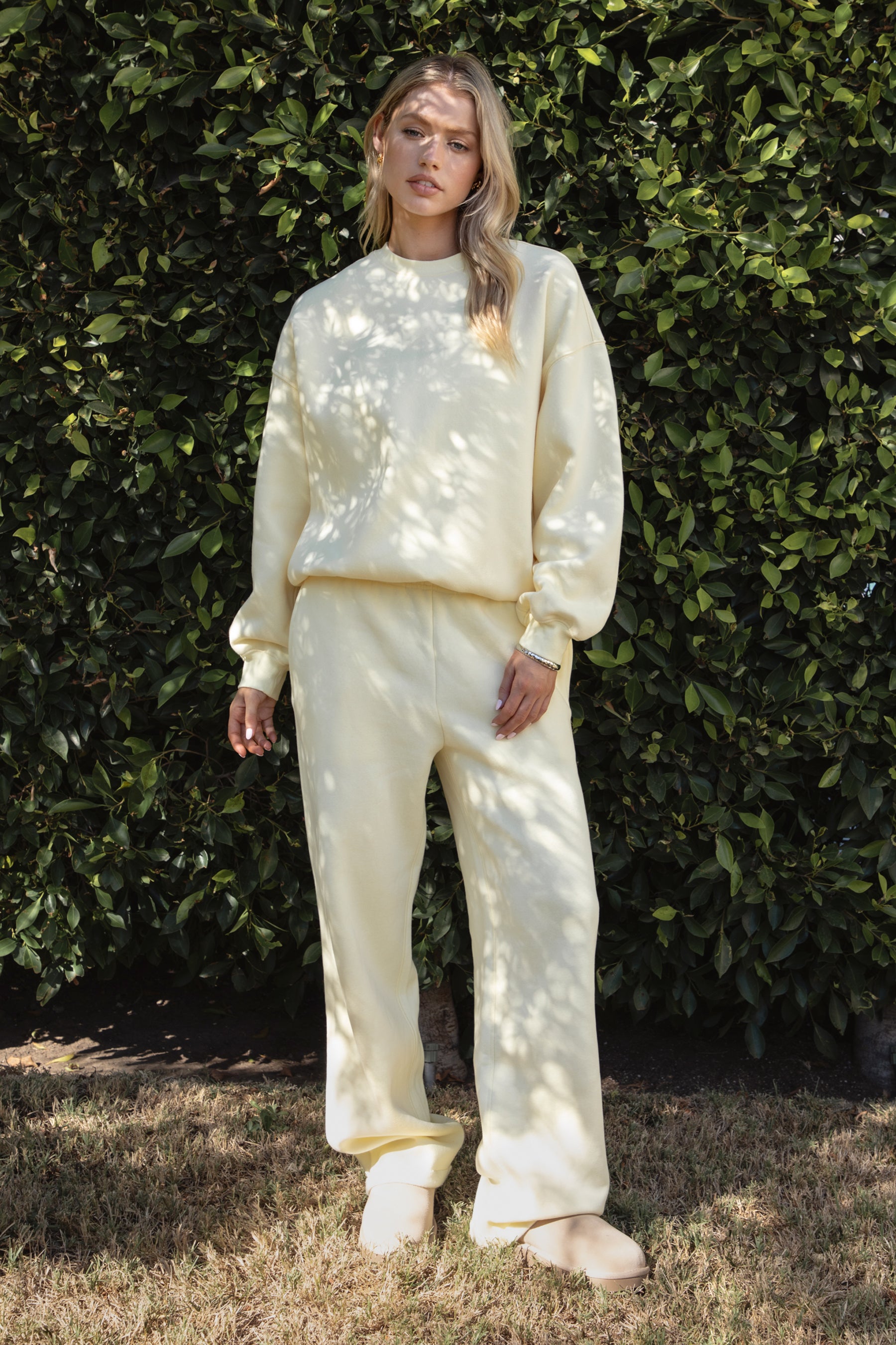 Woman wearing the TIC TOC TH28243 VARSITY pale yellow fleece crew neck pullover sweatshirt featuring dropped shoulders, ribbed trim, and plush fleece fabric with the matching bottoms standing against a green hedge.