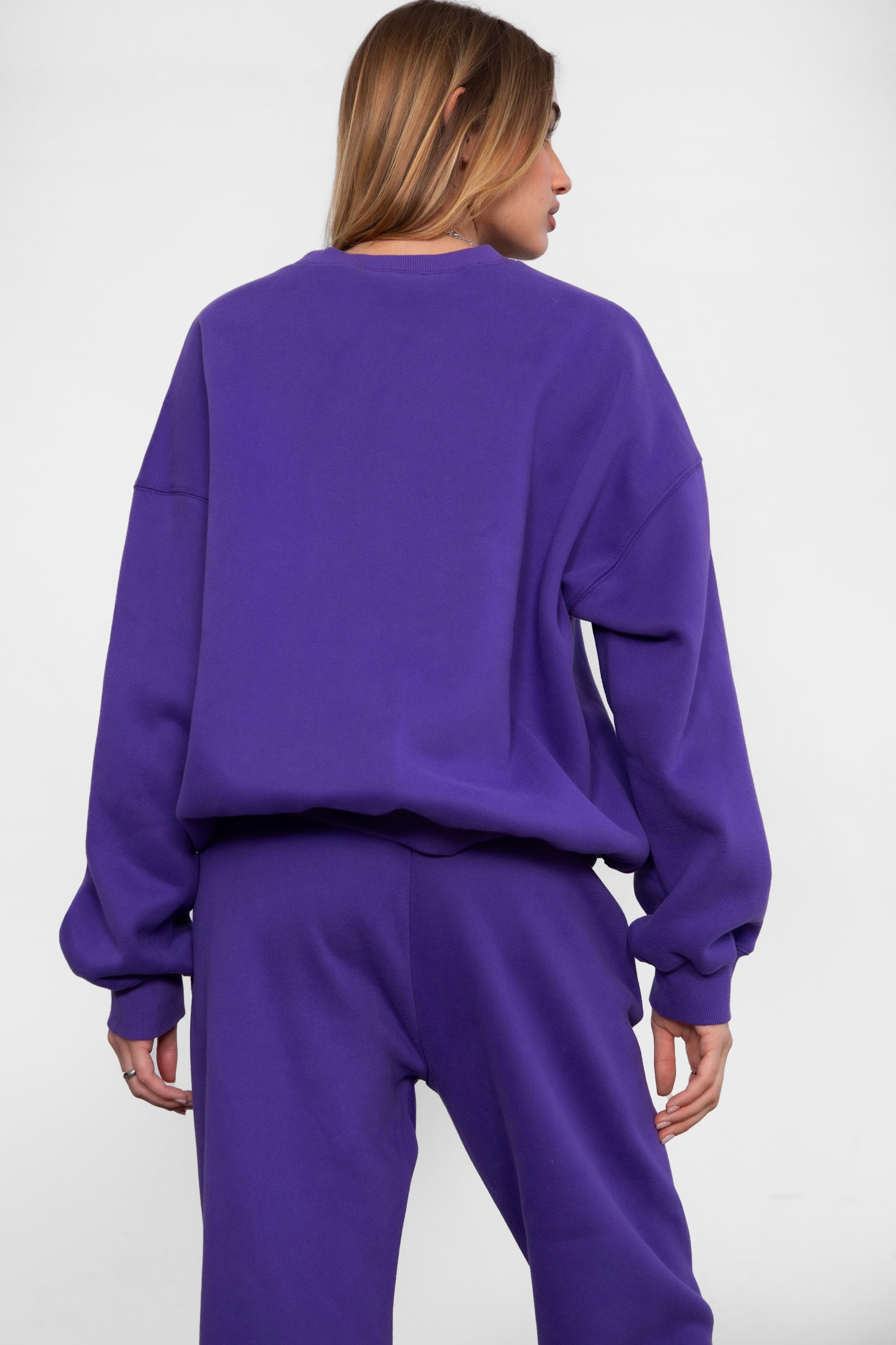 Woman wearing the TIC TOC TH28243 VARSITY purple fleece crew neck pullover sweatshirt featuring dropped shoulders, ribbed trim, and plush fleece fabric with the matching bottoms 