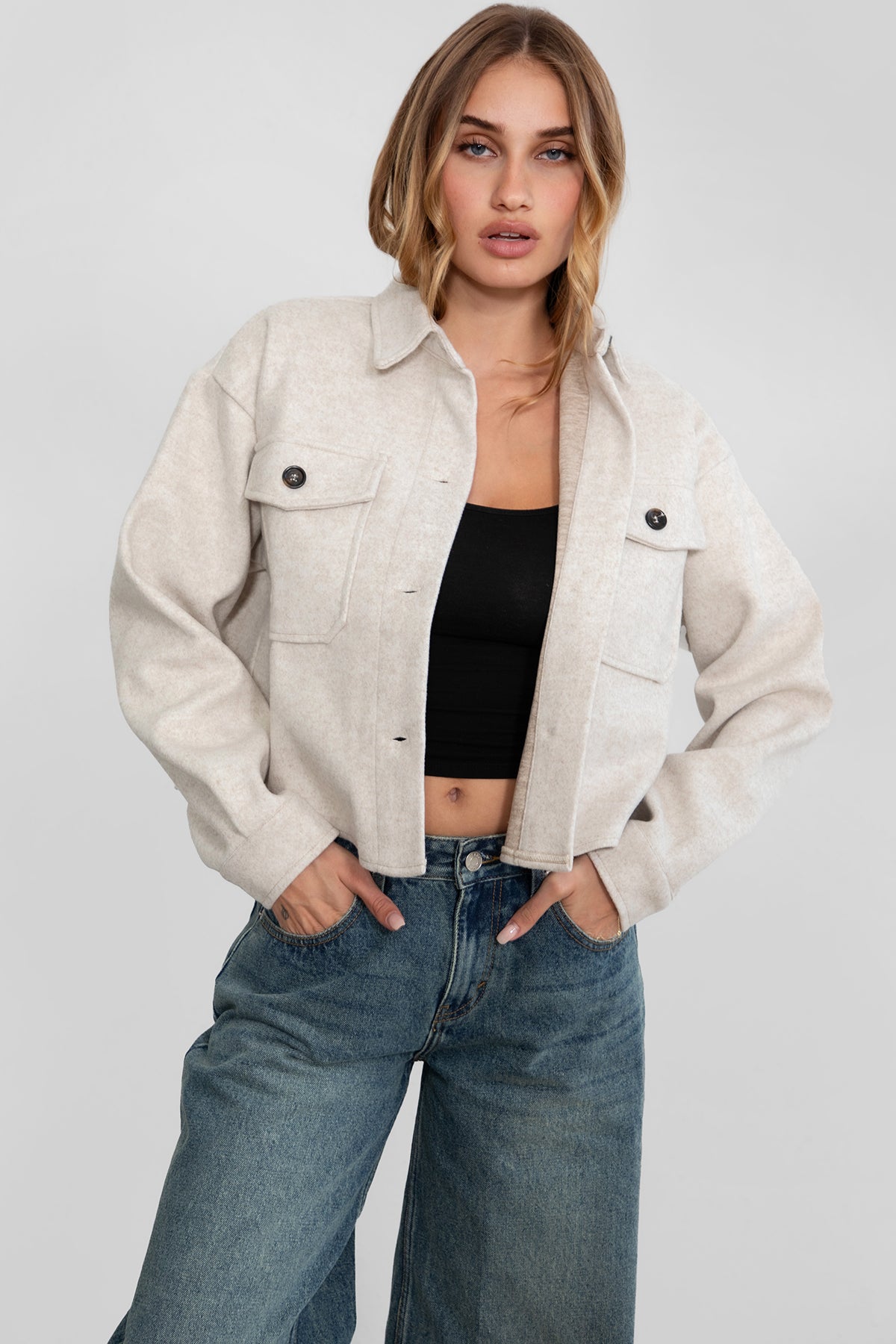 Woman wearing TIC TOC TH29147 WOOLIE faux wool cropped shacket with button front, collared neckline, and double pocket detail in color oatmeal paired with denim pants. 
