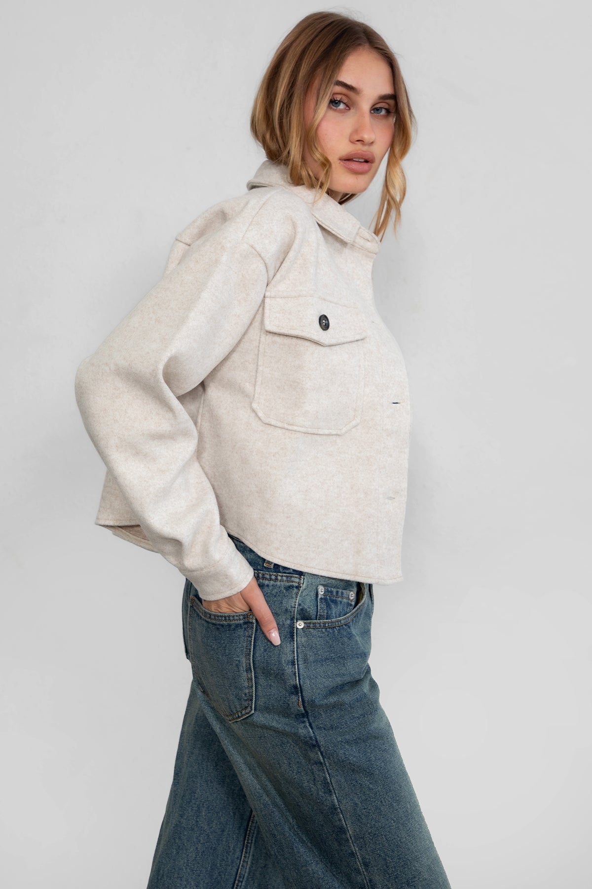 Woman wearing TIC TOC TH29147 WOOLIE faux wool cropped shacket with button front, collared neckline, and double pocket detail in color oatmeal paired with denim pants. 