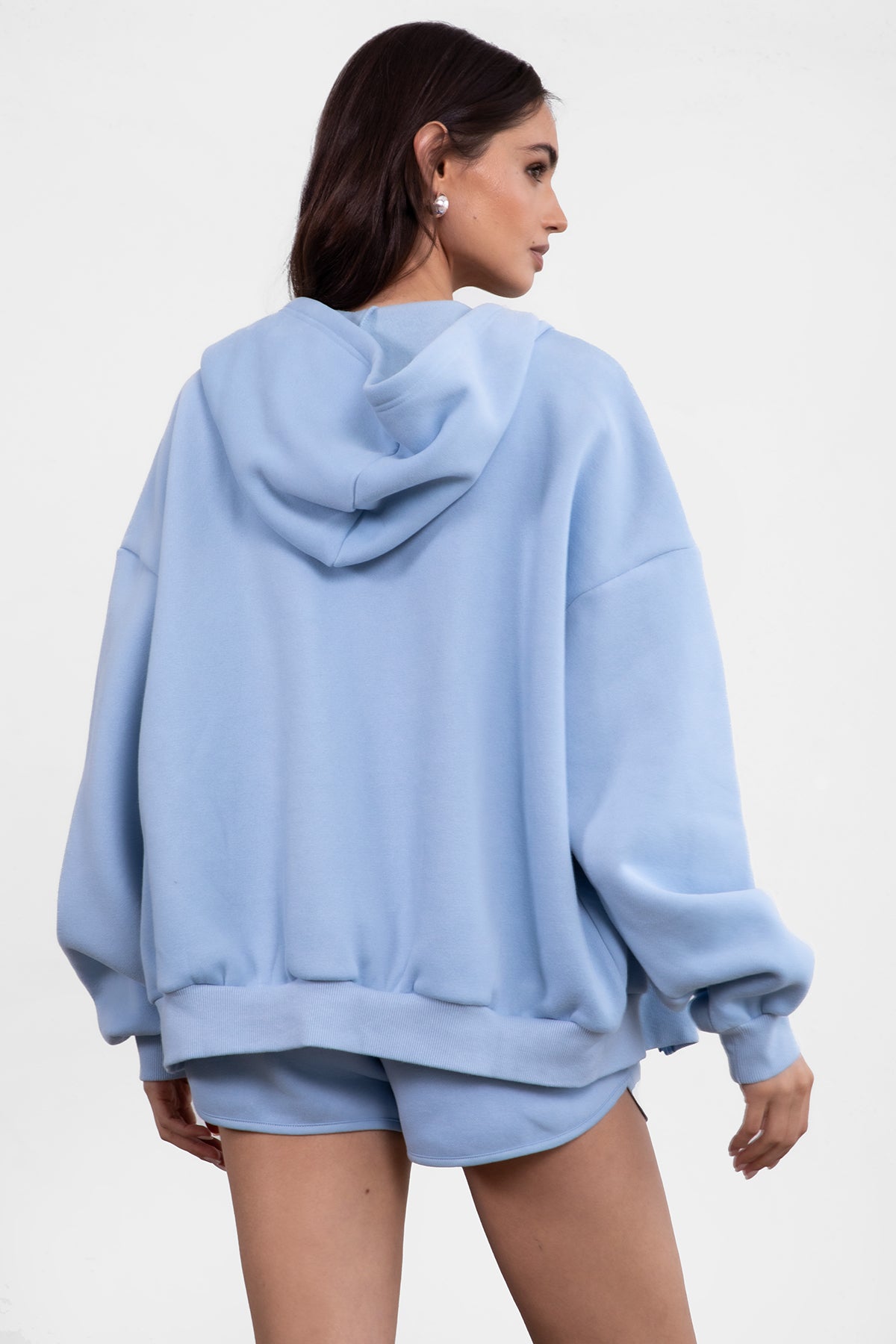 Woman wearing the TIC TOC TH29489 UNIVERSITY baby blue oversized loose fit zip up hoodie featuring dropped shoulders, ribbed hem, and plush fleece fabric with matching bottoms standing against a grey background.