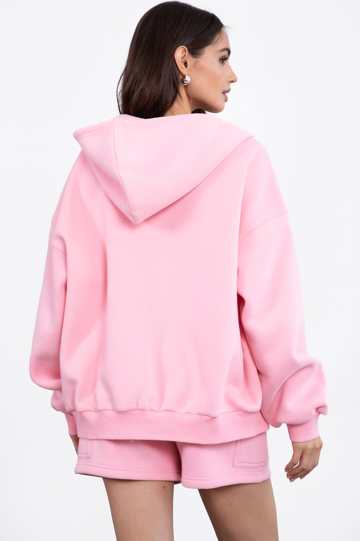 Woman wearing the TIC TOC TH29489 UNIVERSITY blush oversized loose fit zip up hoodie featuring dropped shoulders, ribbed hem, and plush fleece fabric with matching bottoms standing against a grey background.