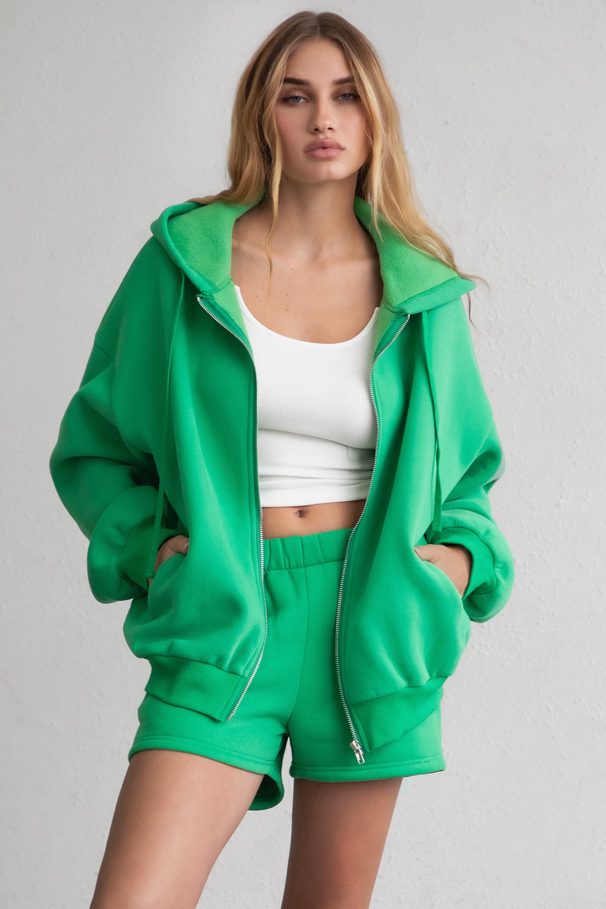 Woman wearing the TIC TOC TH29489 UNIVERSITY bright green oversized loose fit zip up hoodie featuring dropped shoulders, ribbed hem, and plush fleece fabric with matching bottoms standing against a grey background.