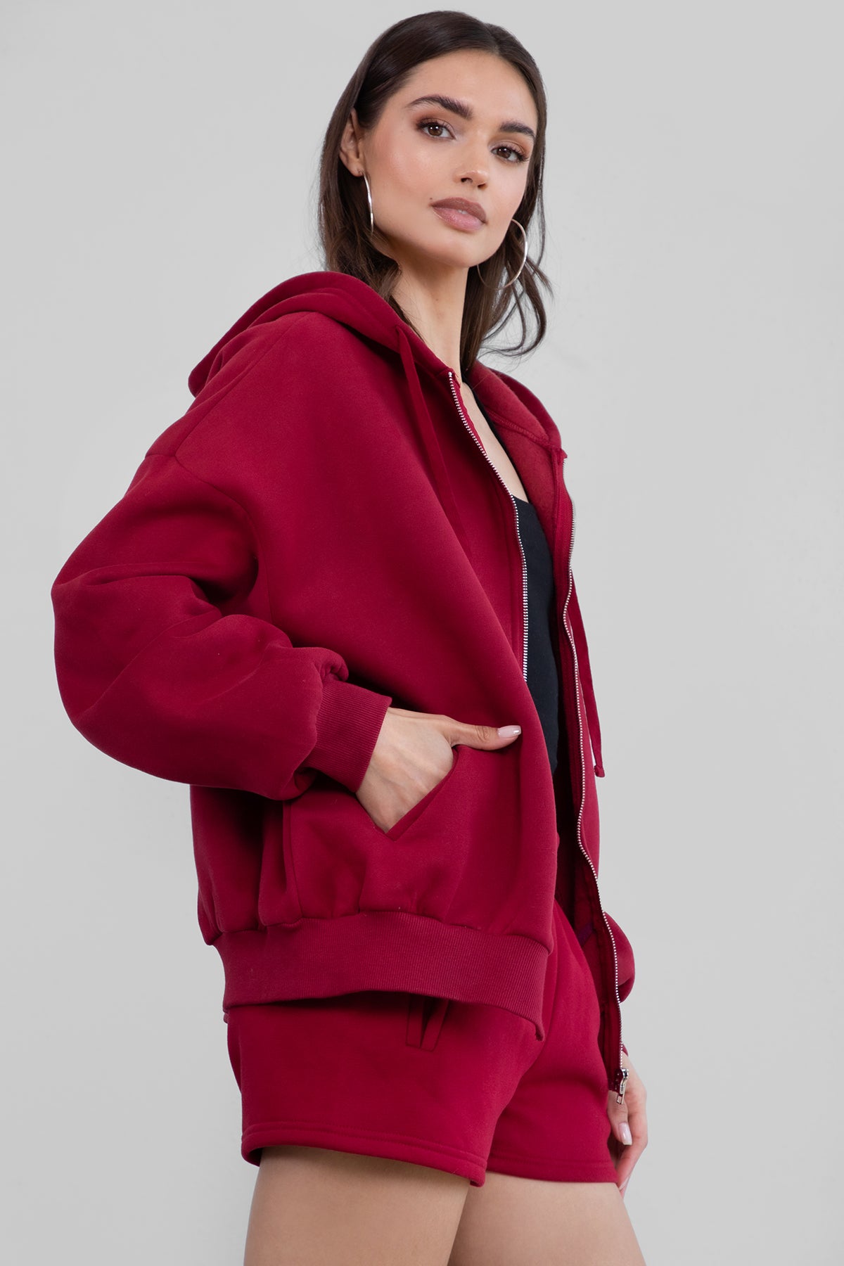 Woman wearing the TIC TOC TH29489 UNIVERSITY burgundy oversized loose fit zip up hoodie featuring dropped shoulders, ribbed hem, and plush fleece fabric with matching bottoms standing against a grey background.