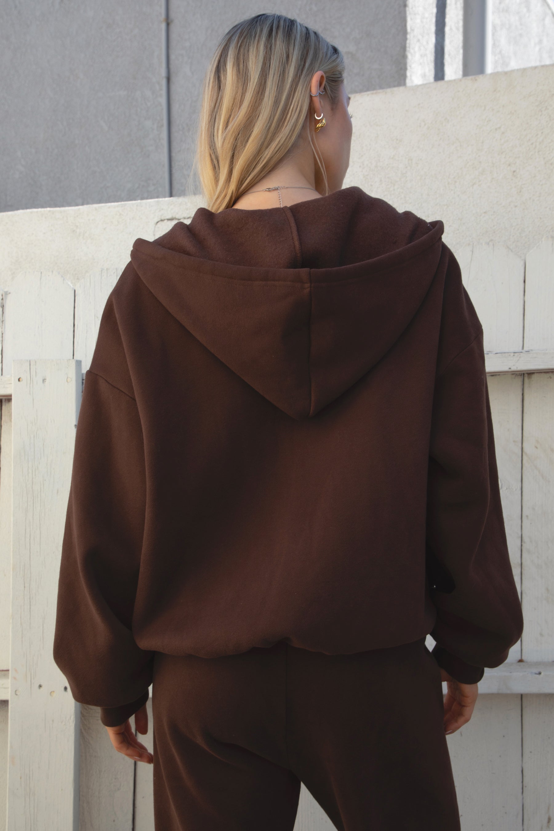 Woman wearing the TIC TOC TH29489 UNIVERSITY brown oversized loose fit zip up hoodie featuring dropped shoulders, ribbed hem, and plush fleece fabric with matching bottoms against a light-colored wall.