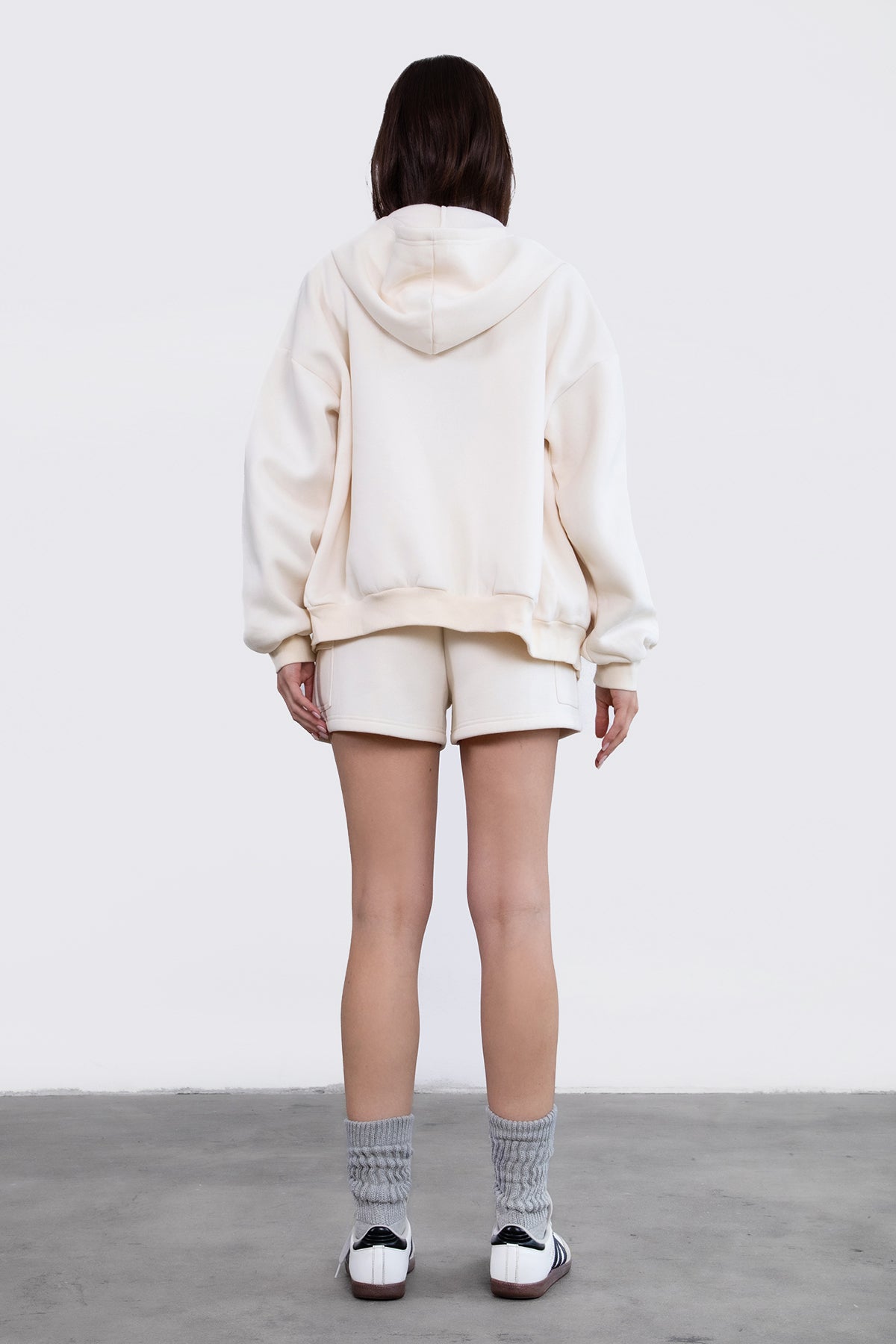 Woman wearing the TIC TOC TH29489 UNIVERSITY ecru oversized loose fit zip up hoodie featuring dropped shoulders, ribbed hem, and plush fleece fabric with matching bottoms standing against a grey background.