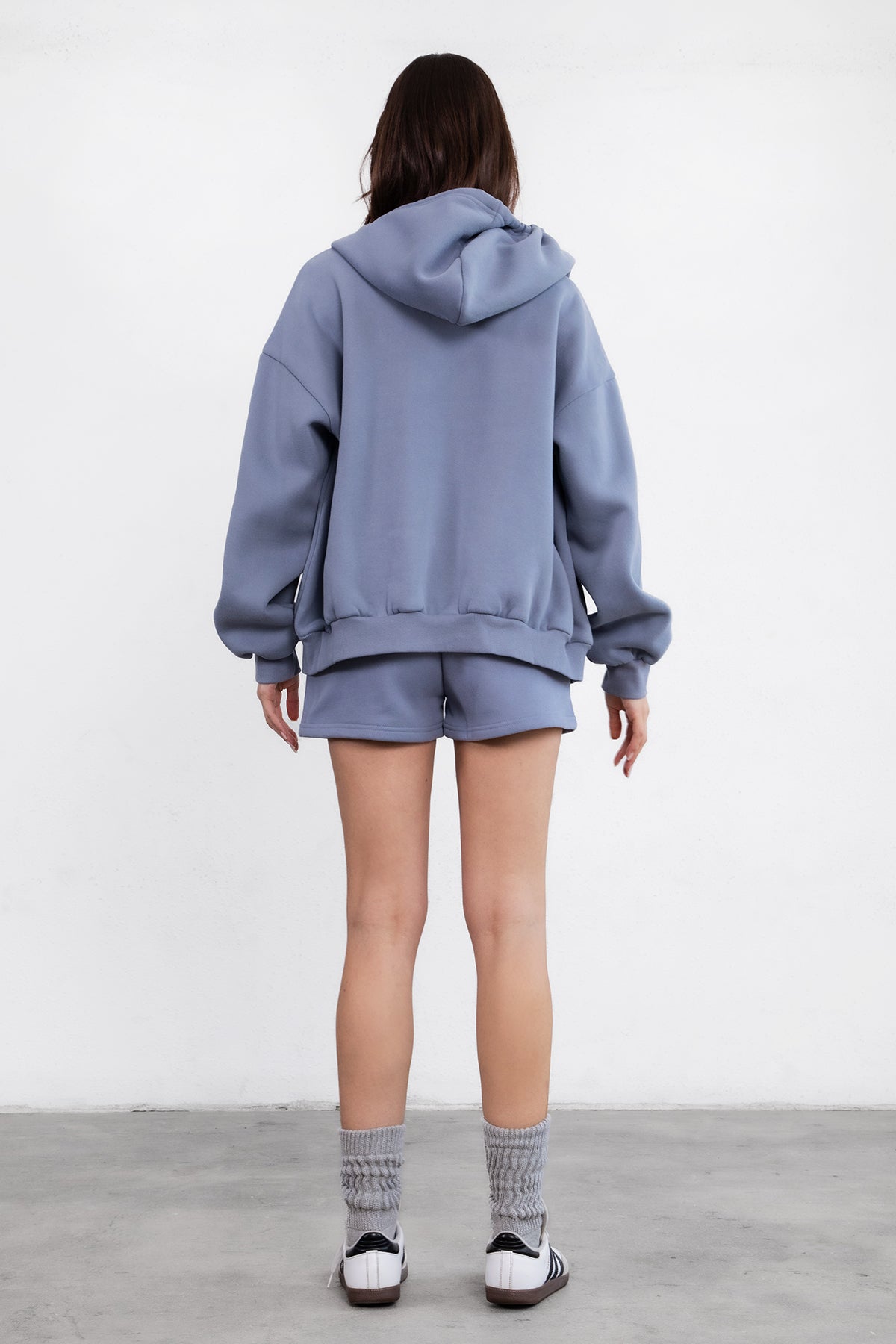Woman wearing the TIC TOC TH29489 UNIVERSITY french blue oversized loose fit zip up hoodie featuring dropped shoulders, ribbed hem, and plush fleece fabric with matching bottoms standing against a grey background.