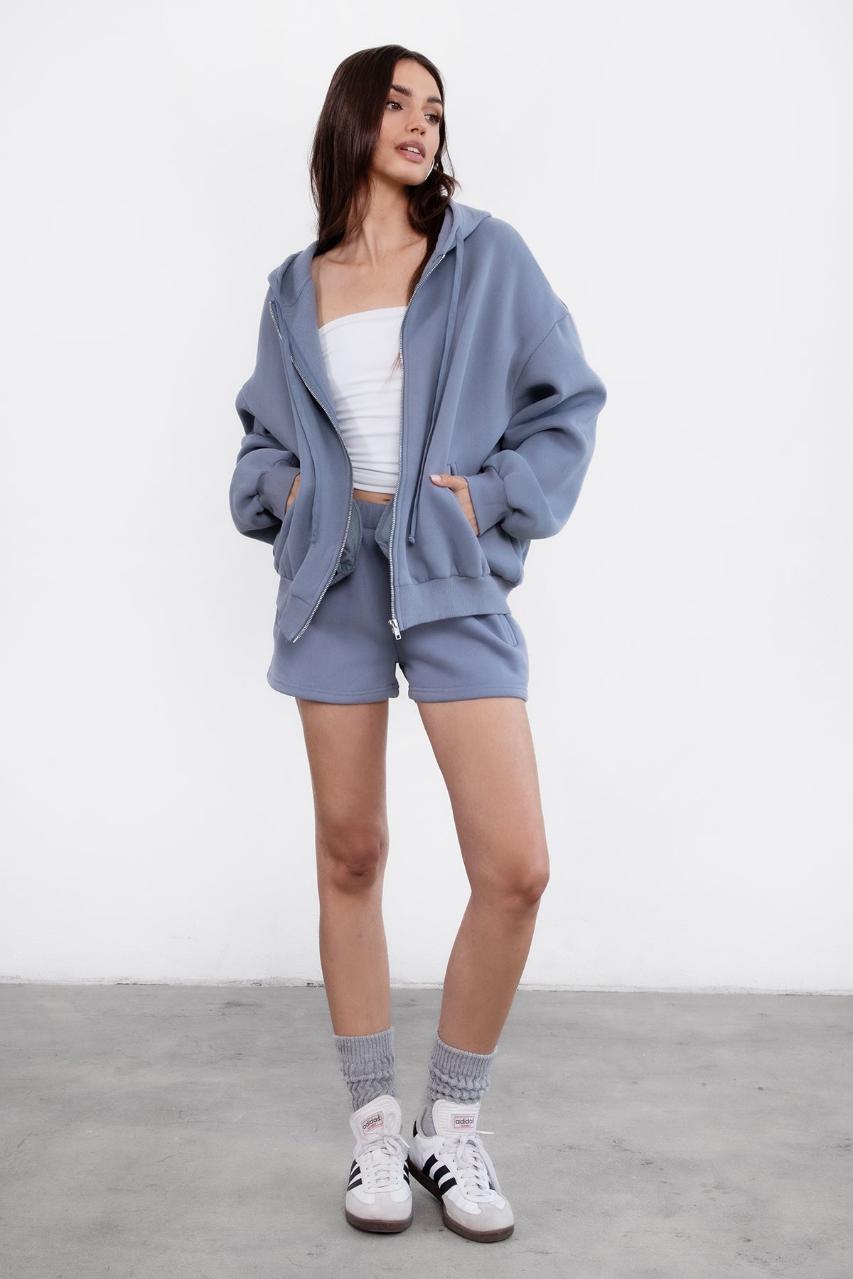 Woman wearing the TIC TOC TH29489 UNIVERSITY french blue oversized loose fit zip up hoodie featuring dropped shoulders, ribbed hem, and plush fleece fabric with matching bottoms standing against a grey background.