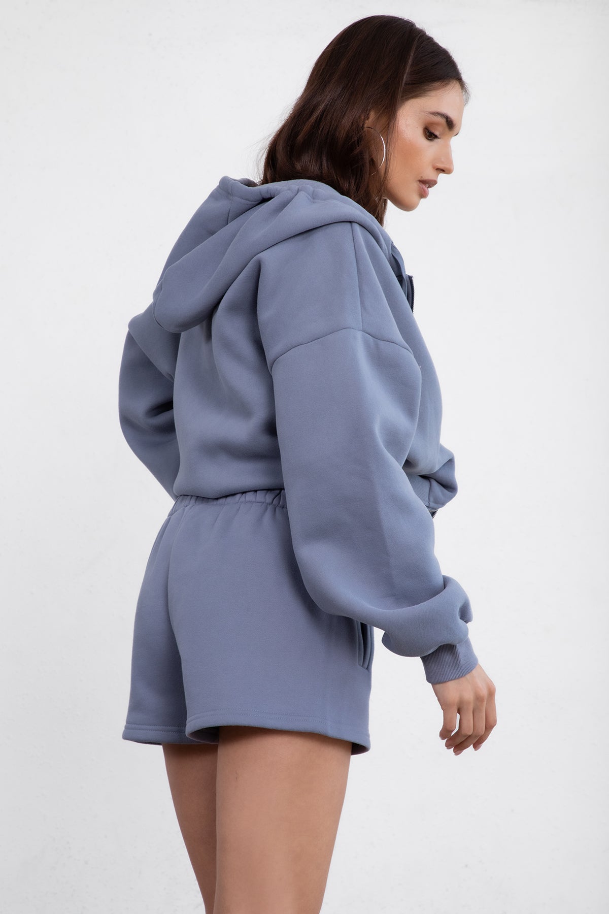 Woman wearing the TIC TOC TH29489 UNIVERSITY french blue oversized loose fit zip up hoodie featuring dropped shoulders, ribbed hem, and plush fleece fabric with matching bottoms standing against a grey background.