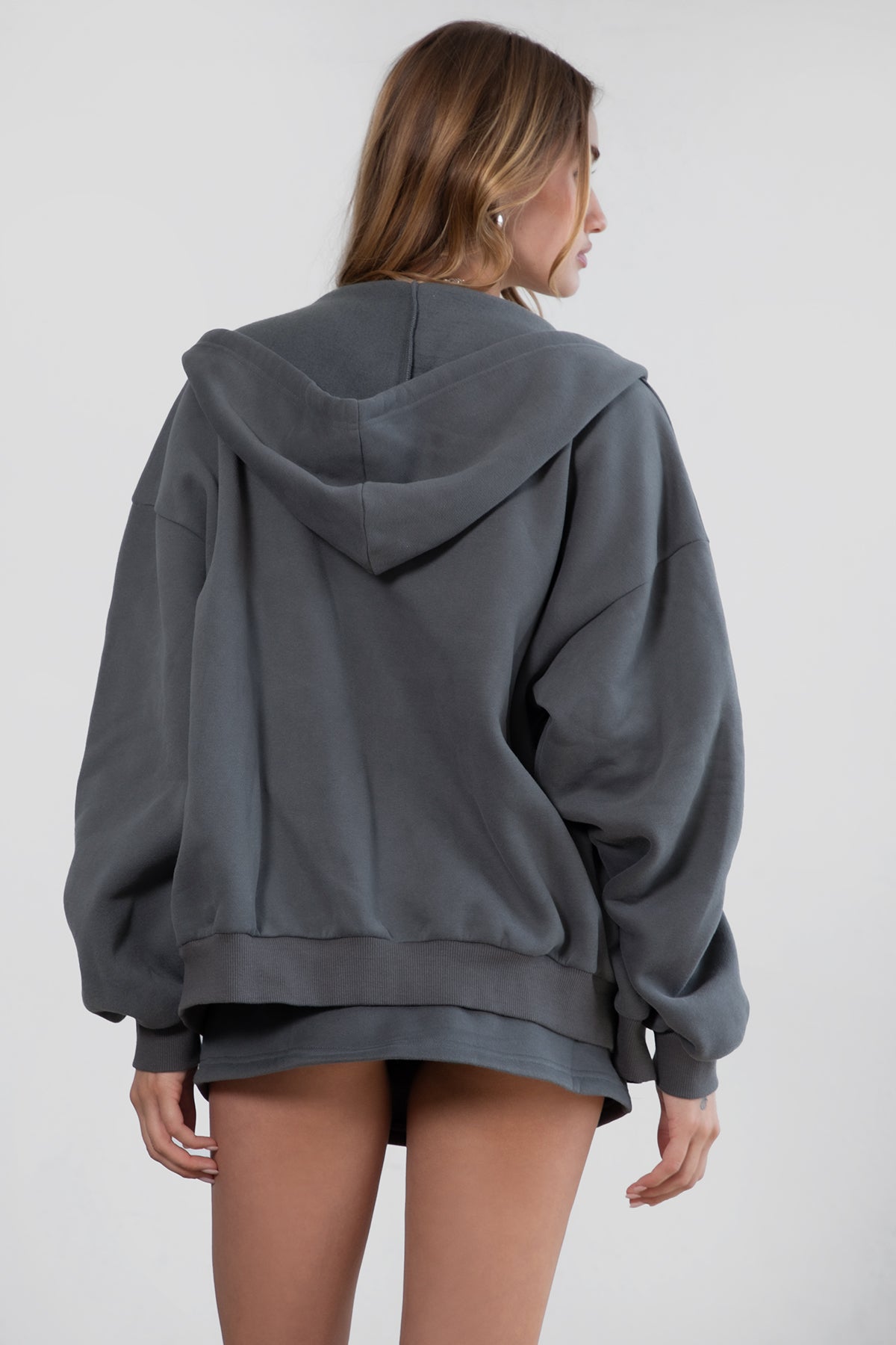Woman wearing the TIC TOC TH29489 UNIVERSITY graphite oversized loose fit zip up hoodie featuring dropped shoulders, ribbed hem, and plush fleece fabric with matching bottoms standing against a grey background.