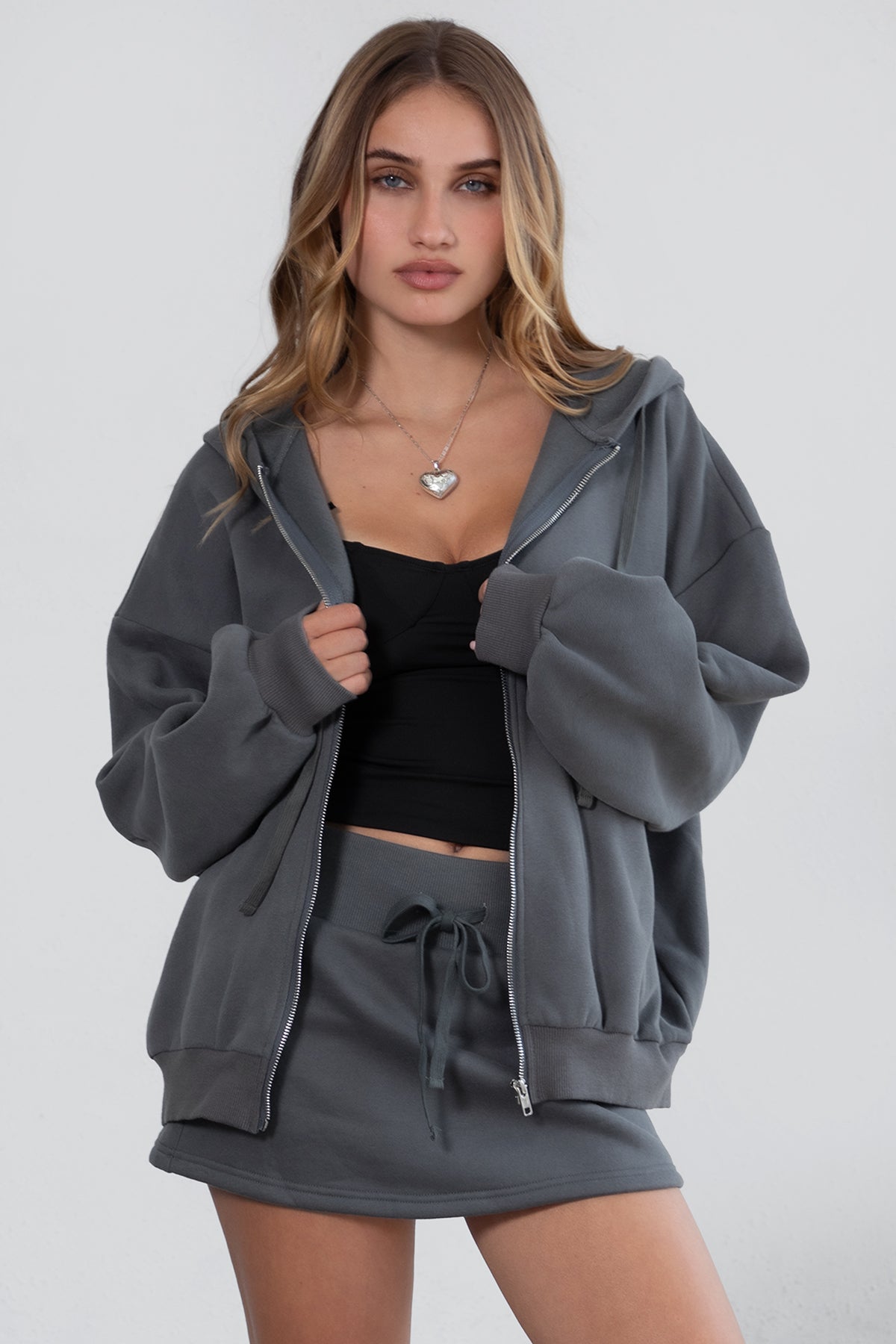 Woman wearing the TIC TOC TH29489 UNIVERSITY graphite oversized loose fit zip up hoodie featuring dropped shoulders, ribbed hem, and plush fleece fabric with matching bottoms standing against a grey background.