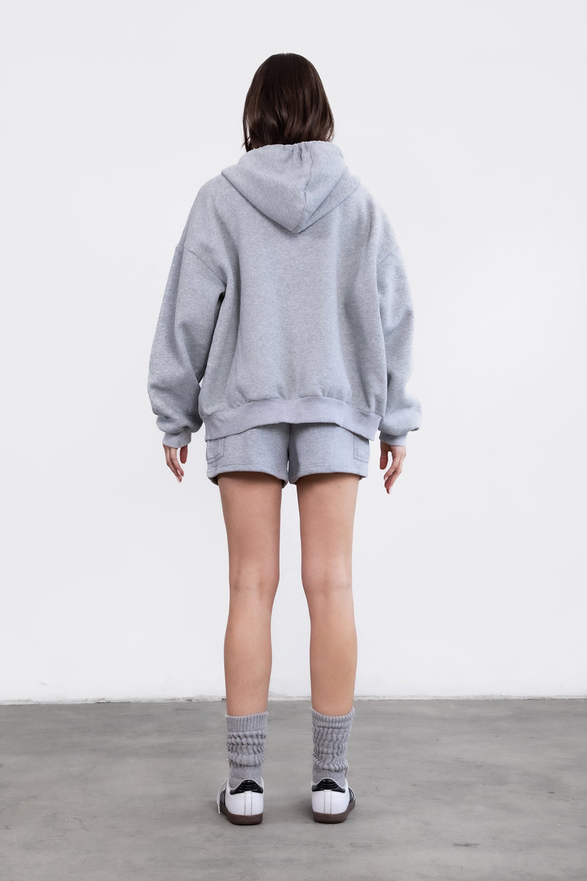 Woman wearing the TIC TOC TH29489 UNIVERSITY heather grey oversized loose fit zip up hoodie featuring dropped shoulders, ribbed hem, and plush fleece fabric with matching bottoms standing against a grey background.