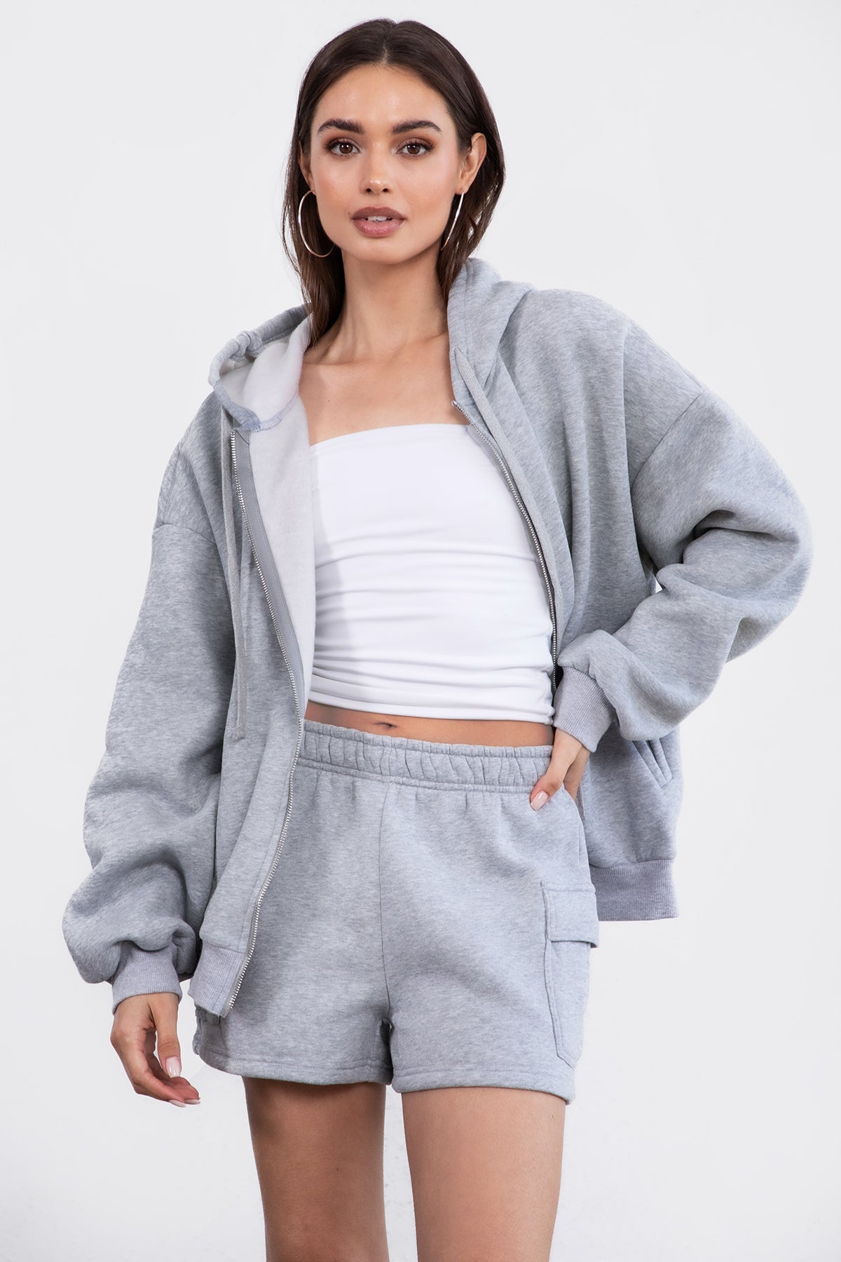 Woman wearing the TIC TOC TH29489 UNIVERSITY heather grey oversized loose fit zip up hoodie featuring dropped shoulders, ribbed hem, and plush fleece fabric with matching bottoms standing against a grey background.