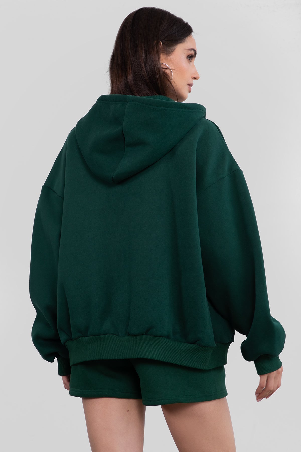 Woman wearing the TIC TOC TH29489 UNIVERSITY hunter green oversized loose fit zip up hoodie featuring dropped shoulders, ribbed hem, and plush fleece fabric with matching bottoms standing against a grey background.