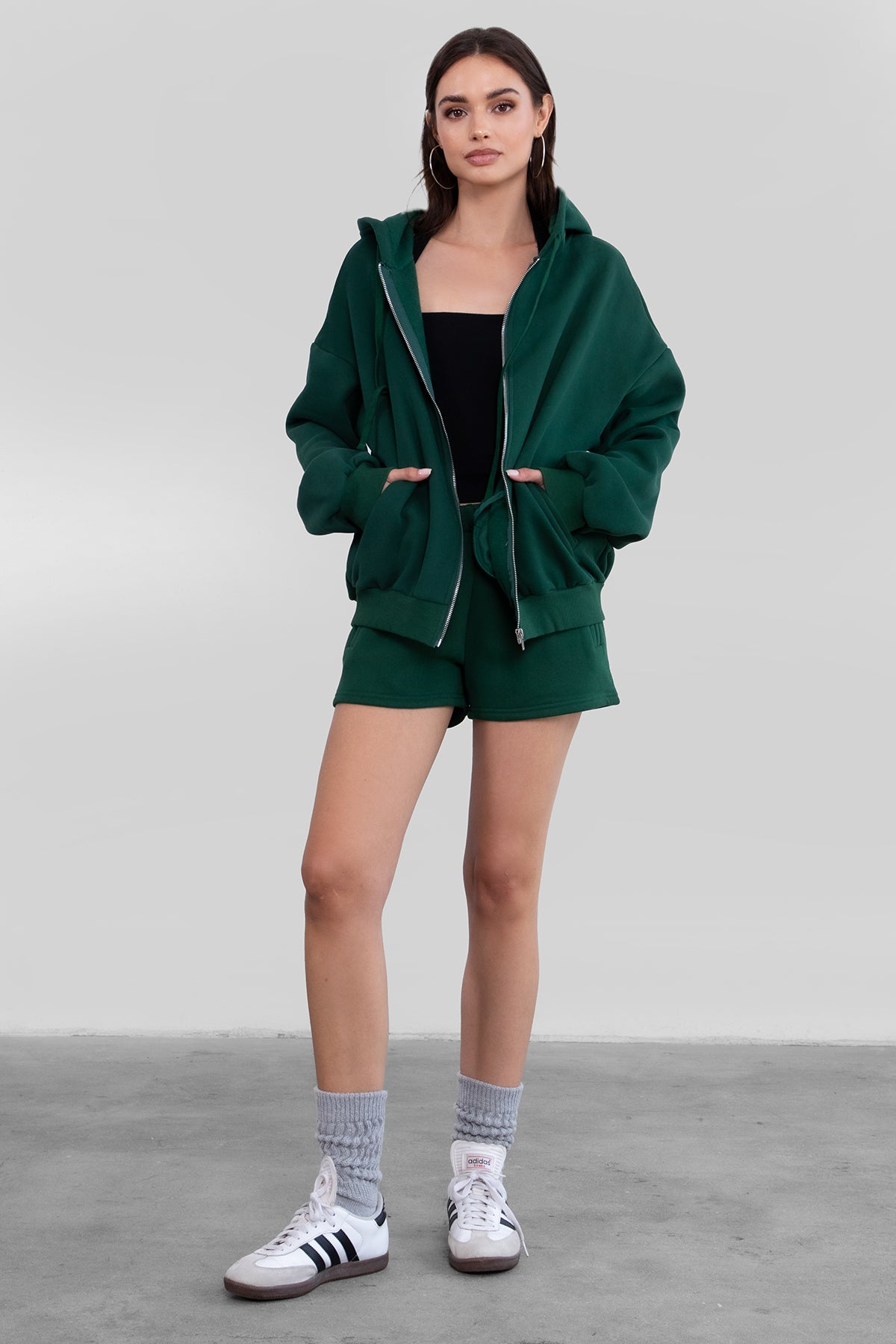 Woman wearing the TIC TOC TH29489 UNIVERSITY hunter green oversized loose fit zip up hoodie featuring dropped shoulders, ribbed hem, and plush fleece fabric with matching bottoms standing against a grey background.