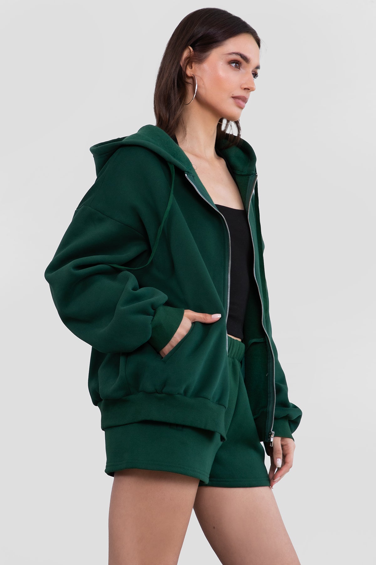 Woman wearing the TIC TOC TH29489 UNIVERSITY hunter green oversized loose fit zip up hoodie featuring dropped shoulders, ribbed hem, and plush fleece fabric with matching bottoms standing against a grey background.