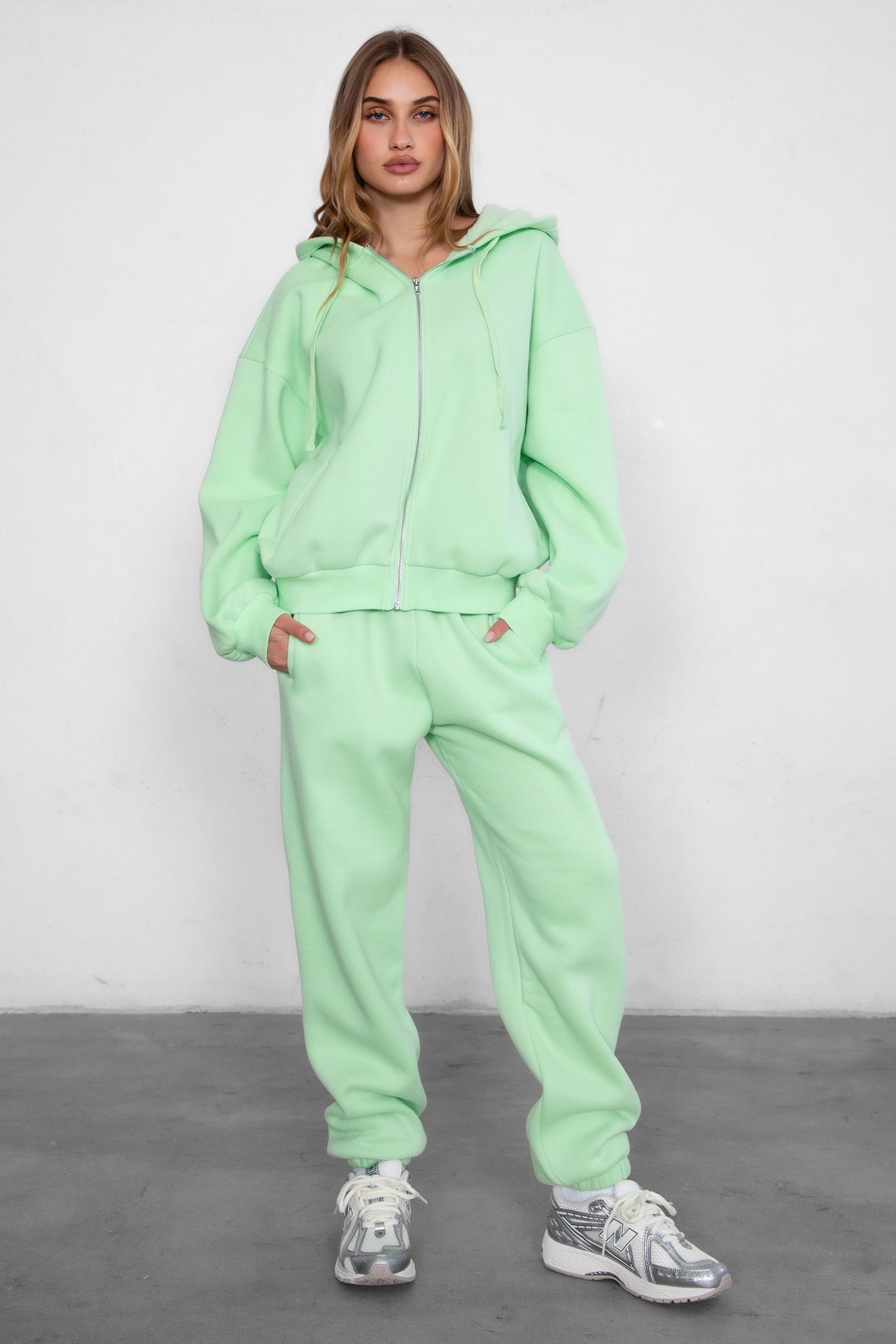Woman wearing the TIC TOC TH29489 UNIVERSITY pastel lime oversized loose fit zip up hoodie featuring dropped shoulders, ribbed hem, and plush fleece fabric with matching bottoms standing against a grey background.