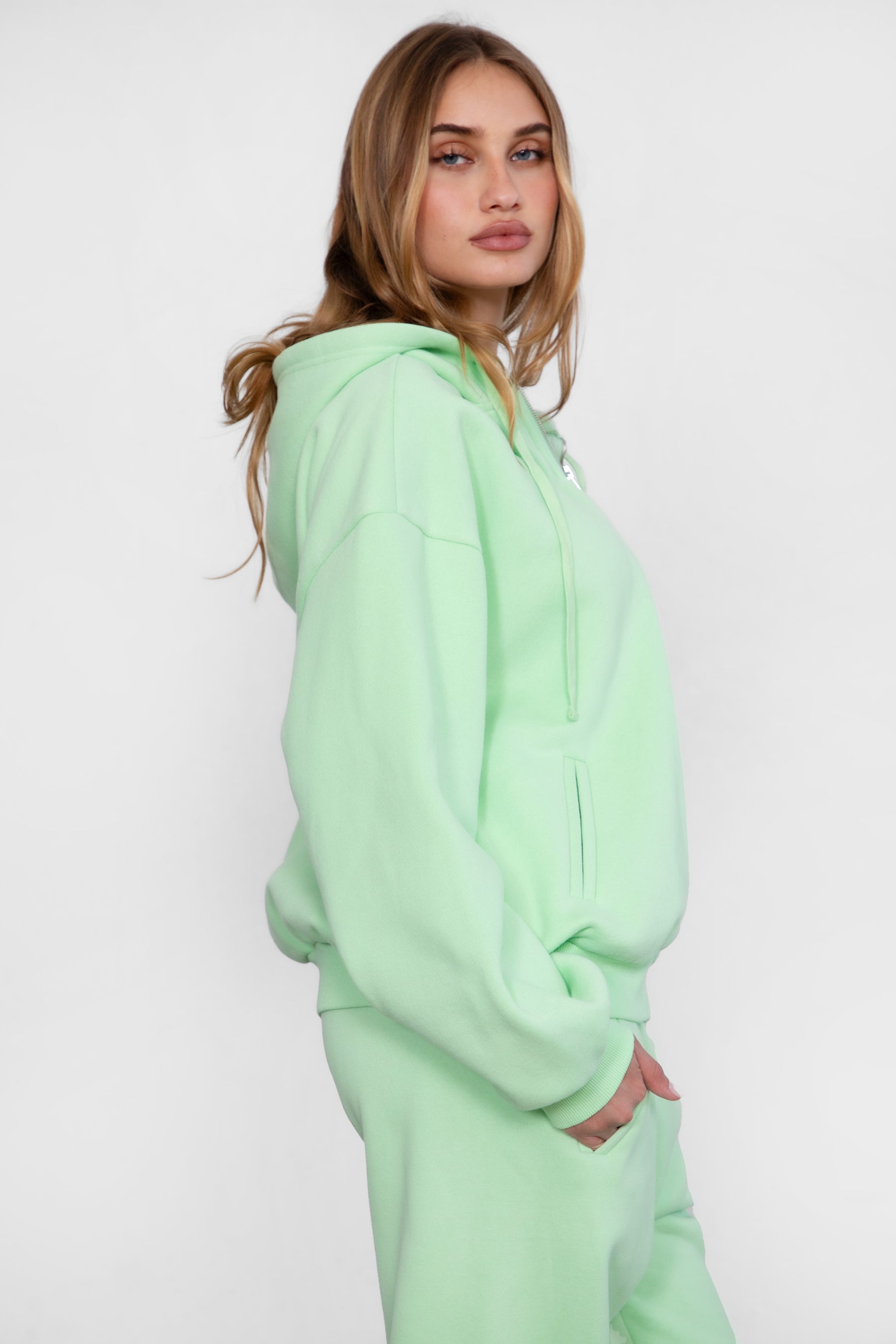 Woman wearing the TIC TOC TH29489 UNIVERSITY pastel lime oversized loose fit zip up hoodie featuring dropped shoulders, ribbed hem, and plush fleece fabric with matching bottoms standing against a grey background.