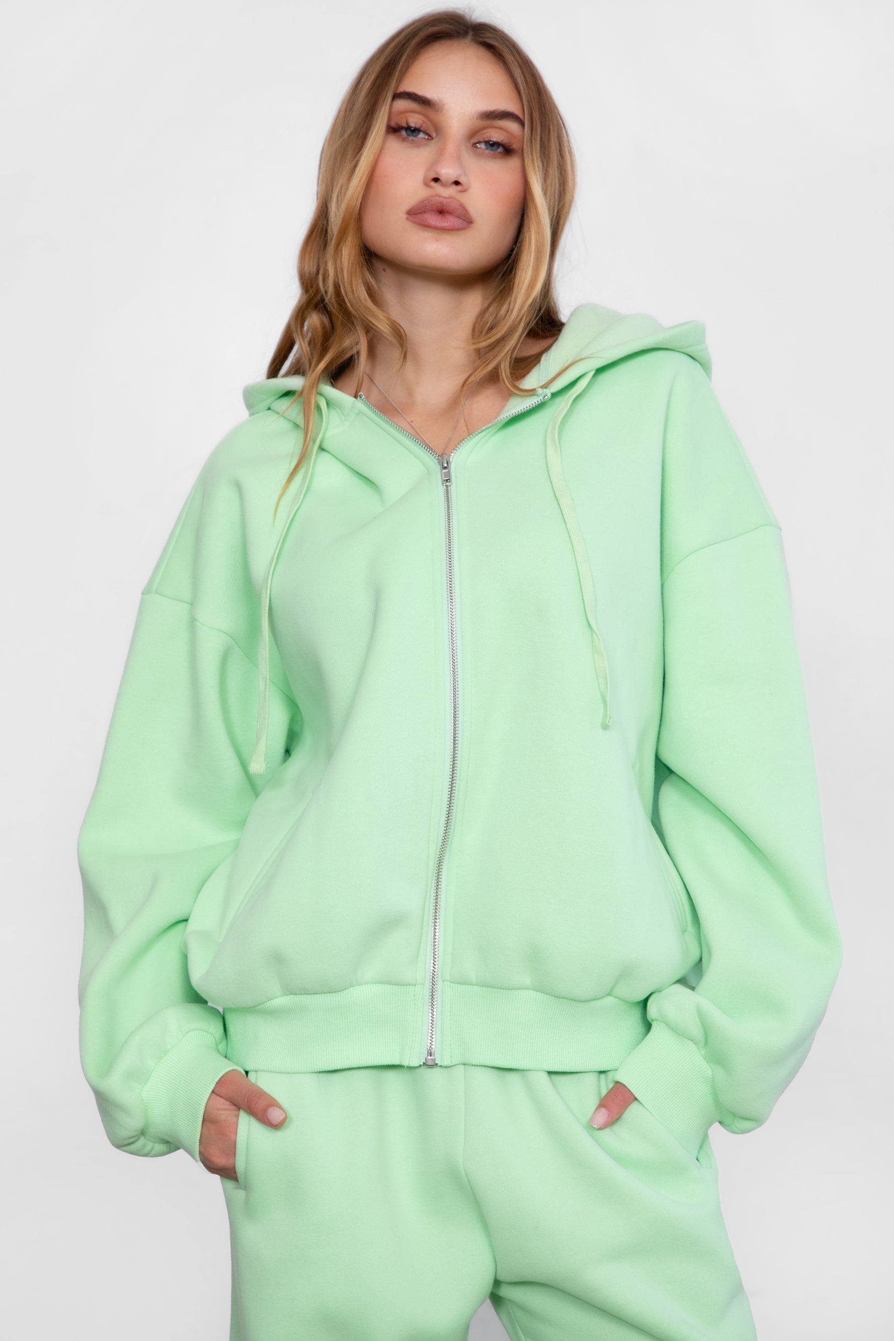 Woman wearing the TIC TOC TH29489 UNIVERSITY pastel lime oversized loose fit zip up hoodie featuring dropped shoulders, ribbed hem, and plush fleece fabric with matching bottoms standing against a grey background.