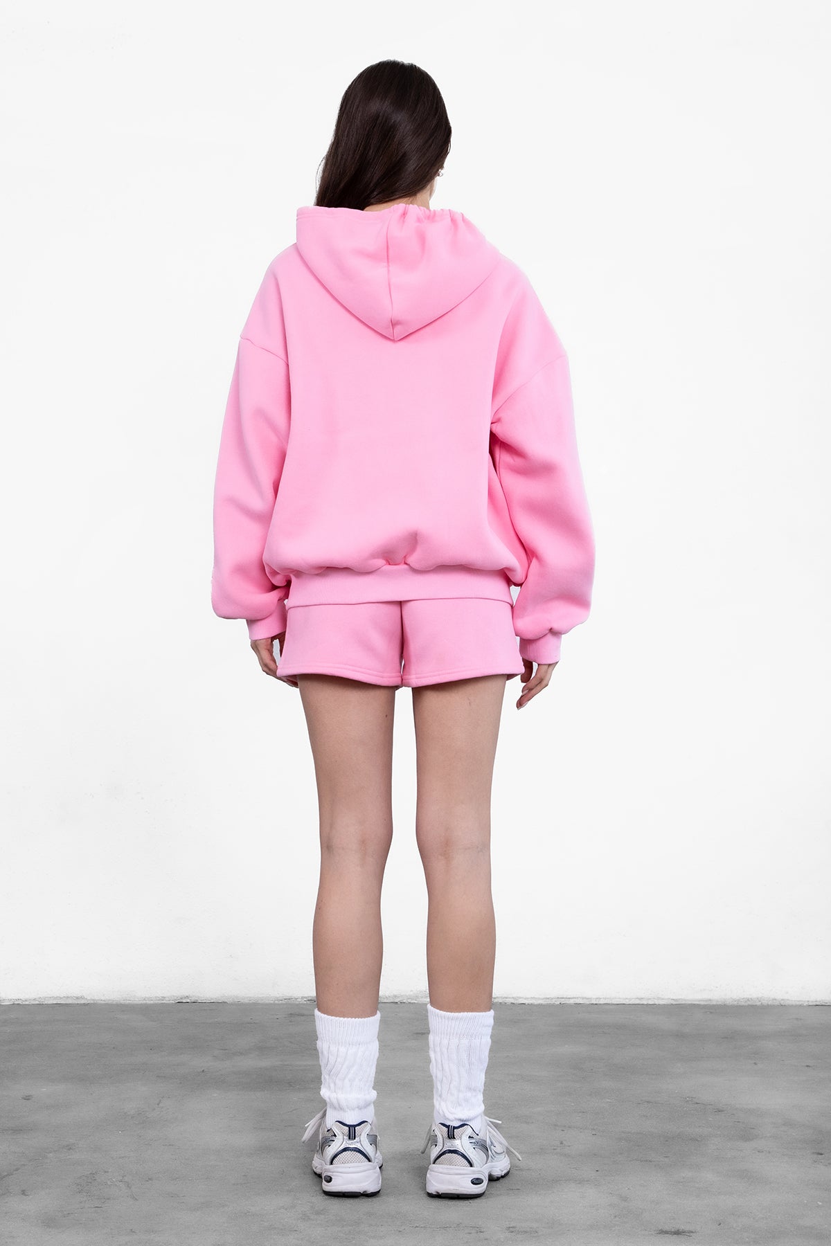 Woman wearing the TIC TOC TH29489 UNIVERSITY pink oversized loose fit zip up hoodie featuring dropped shoulders, ribbed hem, and plush fleece fabric with matching bottoms standing against a grey background.