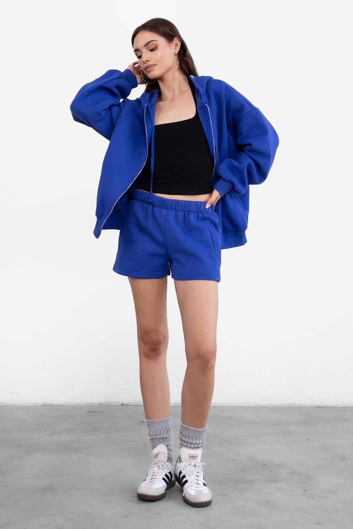 Woman wearing the TIC TOC TH29489 UNIVERSITY royal blue oversized loose fit zip up hoodie featuring dropped shoulders, ribbed hem, and plush fleece fabric with matching bottoms standing against a grey background.