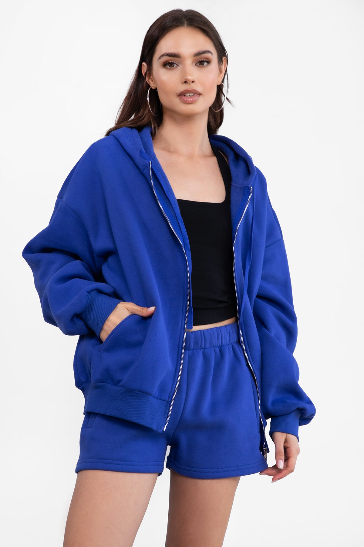Woman wearing the TIC TOC TH29489 UNIVERSITY royal blue oversized loose fit zip up hoodie featuring dropped shoulders, ribbed hem, and plush fleece fabric with matching bottoms standing against a grey background.