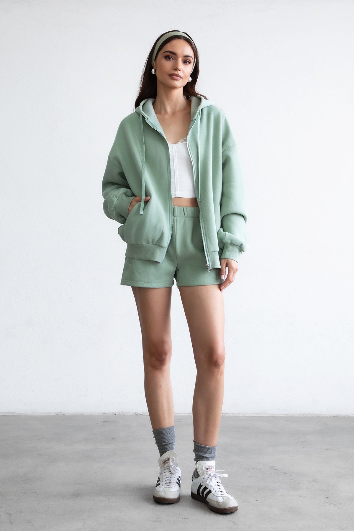 Woman wearing the TIC TOC TH29489 UNIVERSITY sage oversized loose fit zip up hoodie featuring dropped shoulders, ribbed hem, and plush fleece fabric with matching bottoms standing against a grey background.