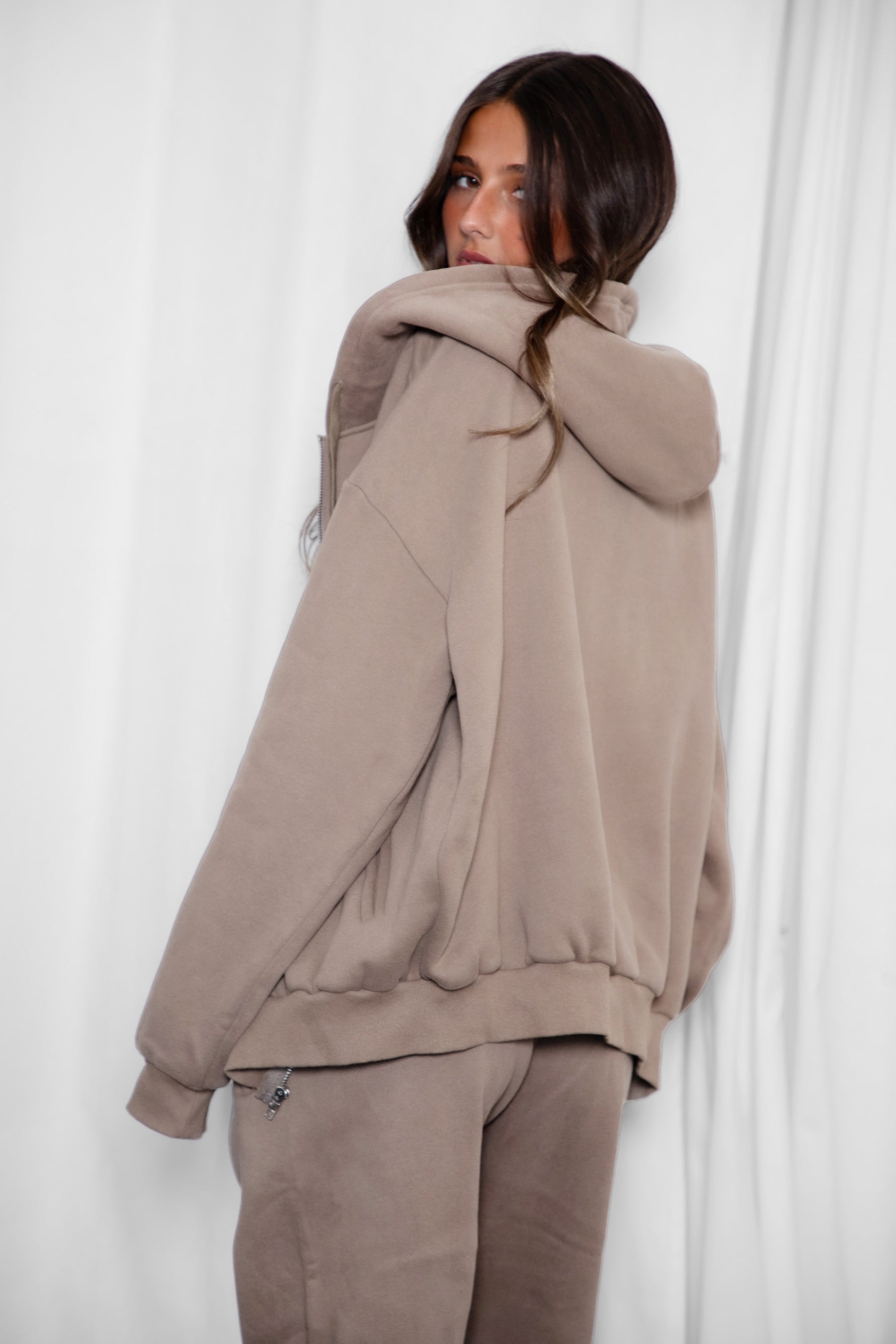 Woman wearing the TIC TOC TH29489 UNIVERSITY taupe oversized loose fit zip up hoodie featuring dropped shoulders, ribbed hem, and plush fleece fabric with matching bottoms standing against a grey background.