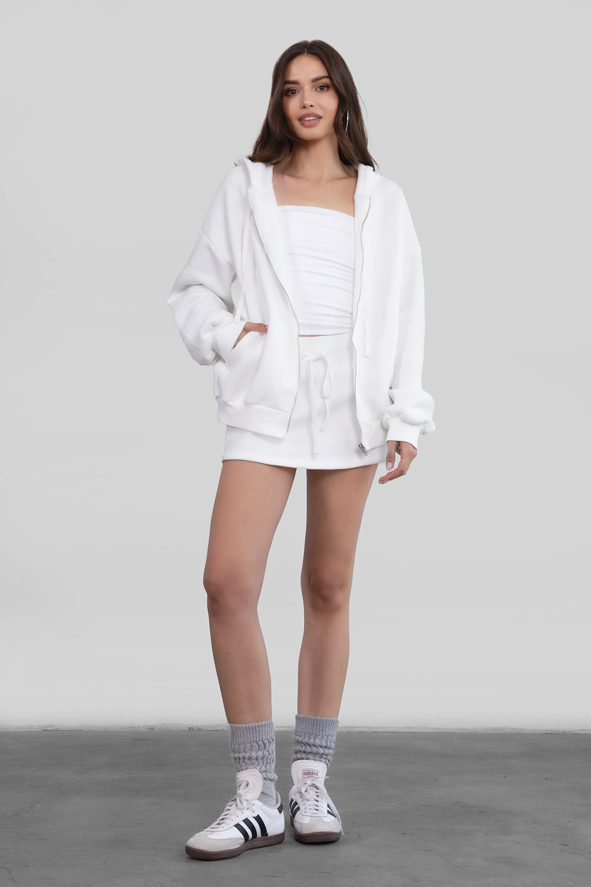 Woman wearing the TIC TOC TH29489 UNIVERSITY white oversized loose fit zip up hoodie featuring dropped shoulders, ribbed hem, and plush fleece fabric with matching bottoms standing against a grey background.