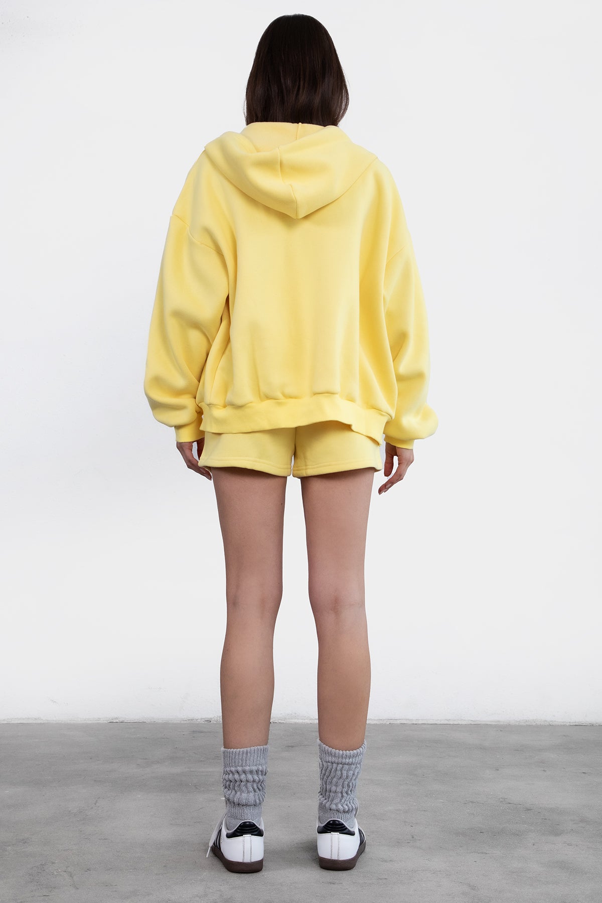 Woman wearing the TIC TOC TH29489 UNIVERSITY yellow oversized loose fit zip up hoodie featuring dropped shoulders, ribbed hem, and plush fleece fabric with matching bottoms standing against a grey background.
