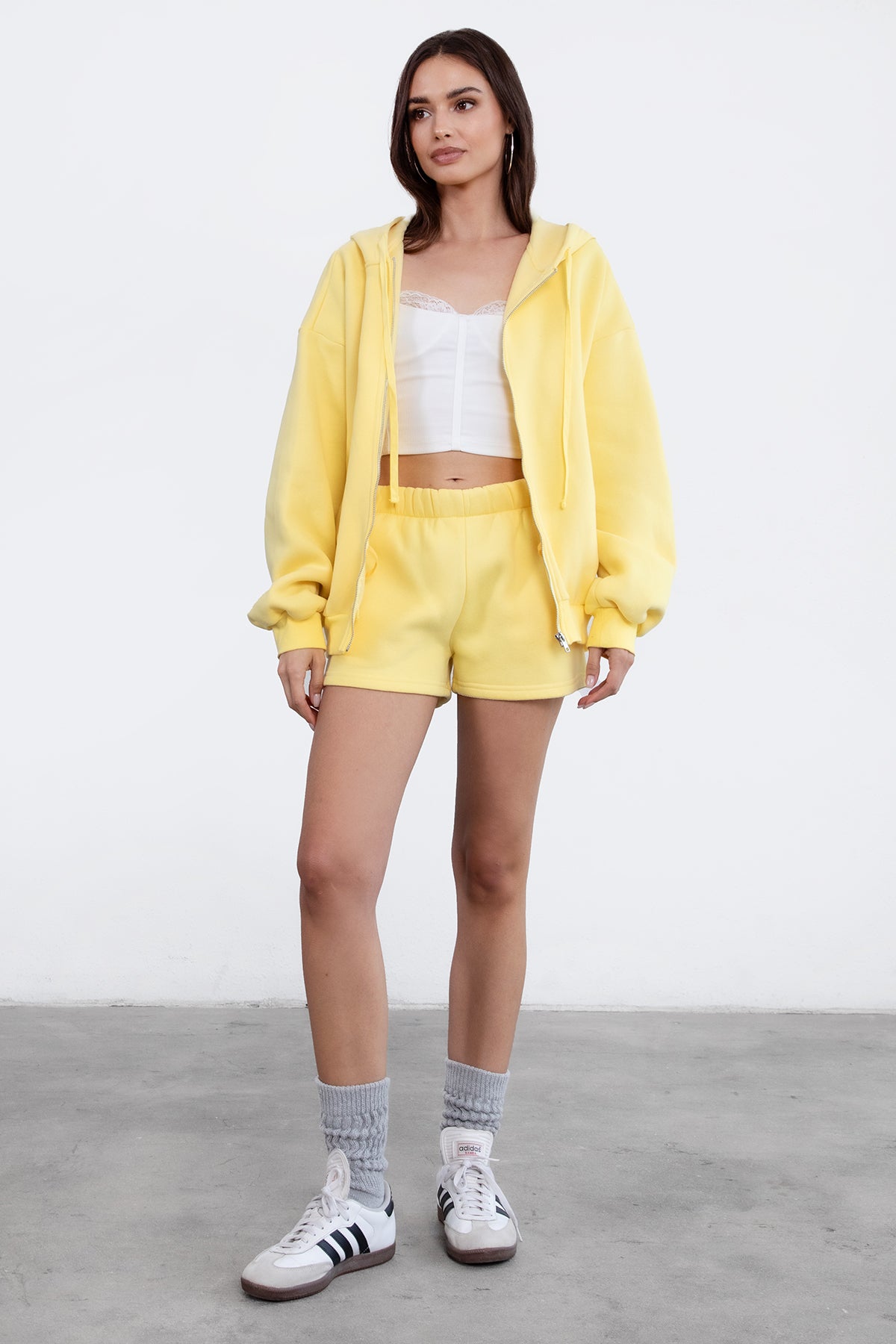 Woman wearing the TIC TOC TH29489 UNIVERSITY yellow oversized loose fit zip up hoodie featuring dropped shoulders, ribbed hem, and plush fleece fabric with matching bottoms standing against a grey background.