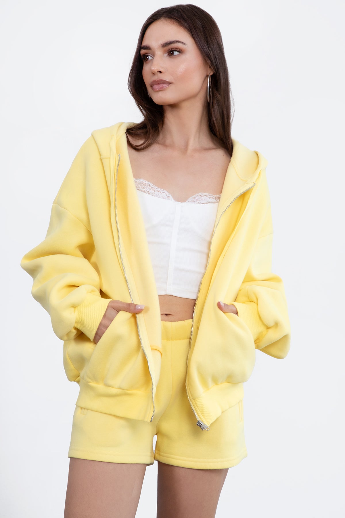 Woman wearing the TIC TOC TH29489 UNIVERSITY yellow oversized loose fit zip up hoodie featuring dropped shoulders, ribbed hem, and plush fleece fabric with matching bottoms standing against a grey background.