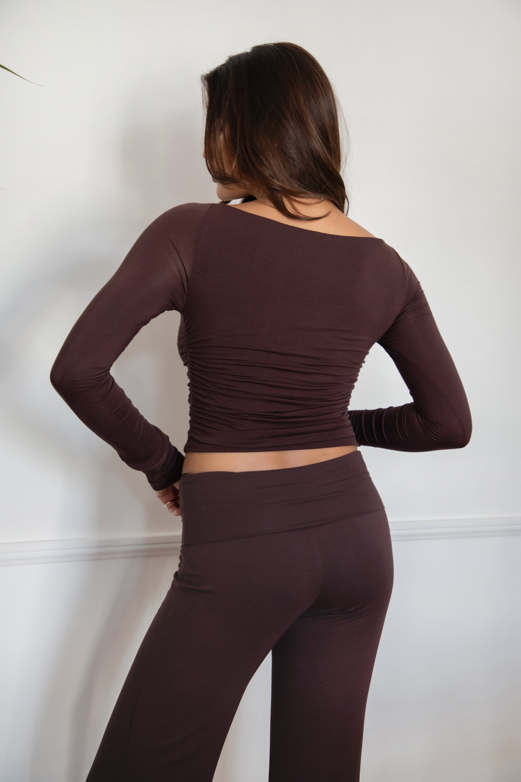 Woman wearing the TIC TOC TH30643 JOLINA brown core basic long sleeve top featuring high boat neckline, thumbhole sleeves, and ruched detailing along the bodice with matching bottoms in an indoor setting.