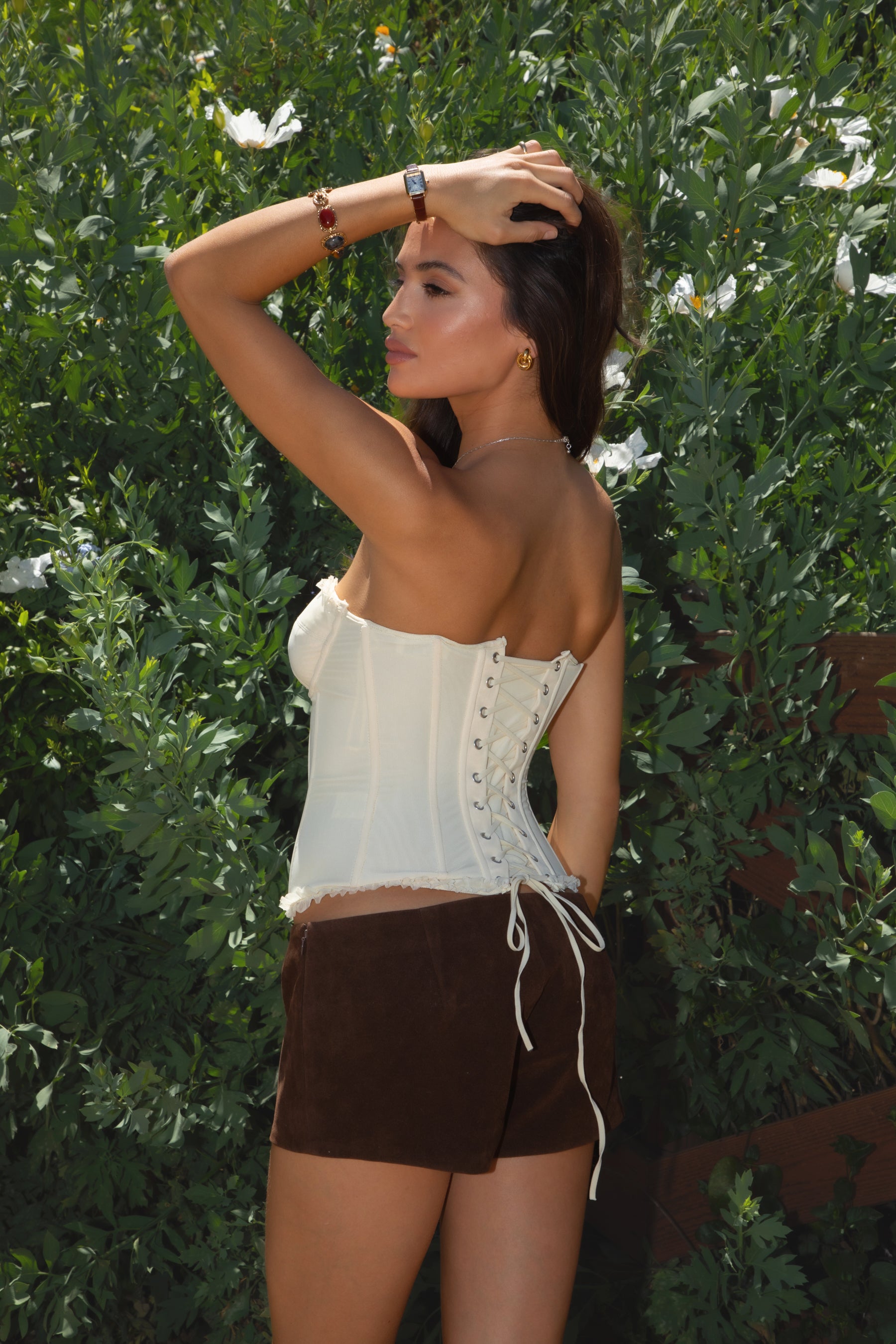 Model wearing the TIC TOC TH30975 LAUREL cream mesh strapless bustier corset top featuring ruffle trim edges, underwire cups, and lace-up back paired with brown suede shorts, standing against  greenery
